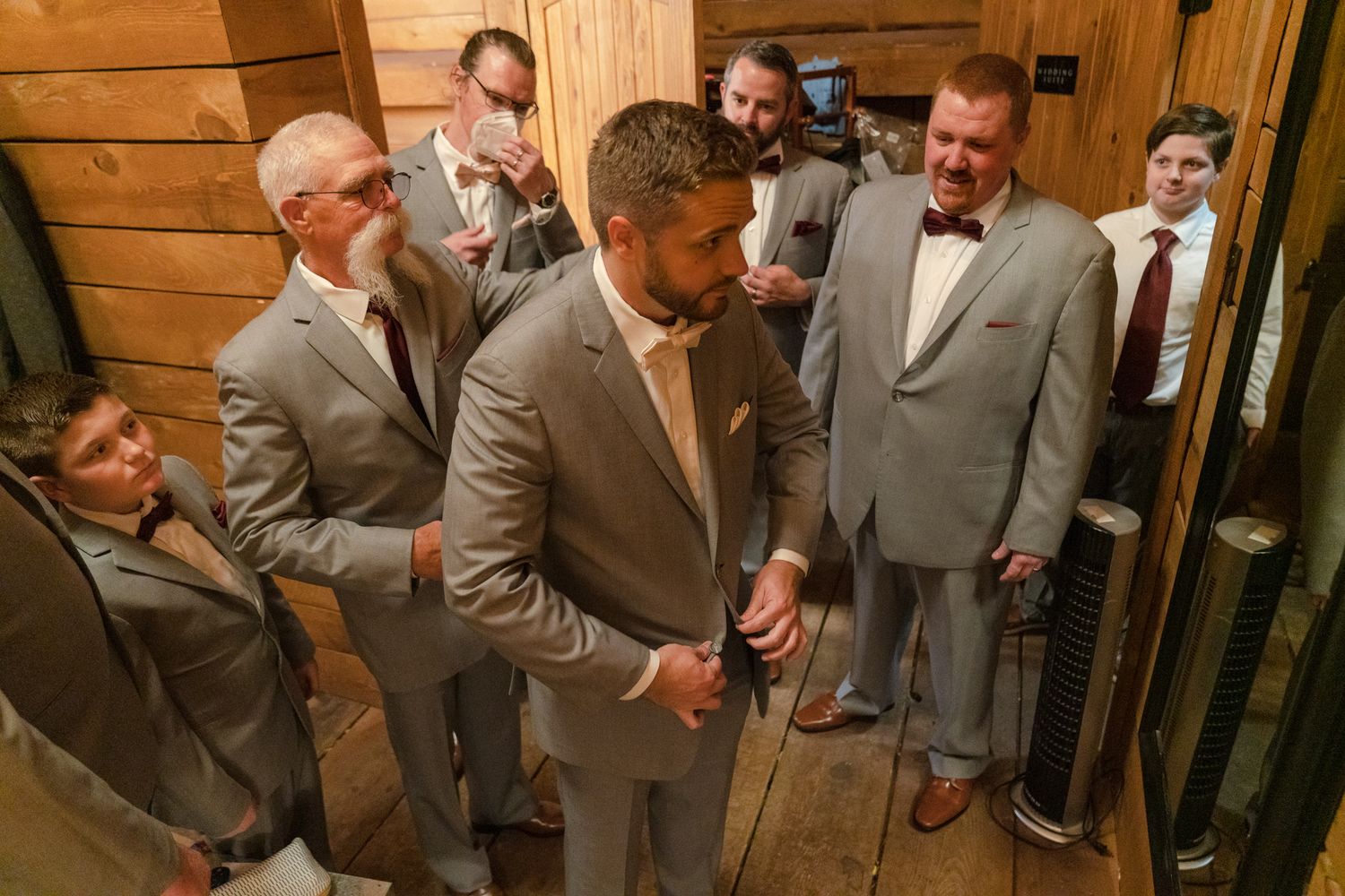 Groomsmen in gray suits gather around in a rustic wooden room before the wedding ceremony.