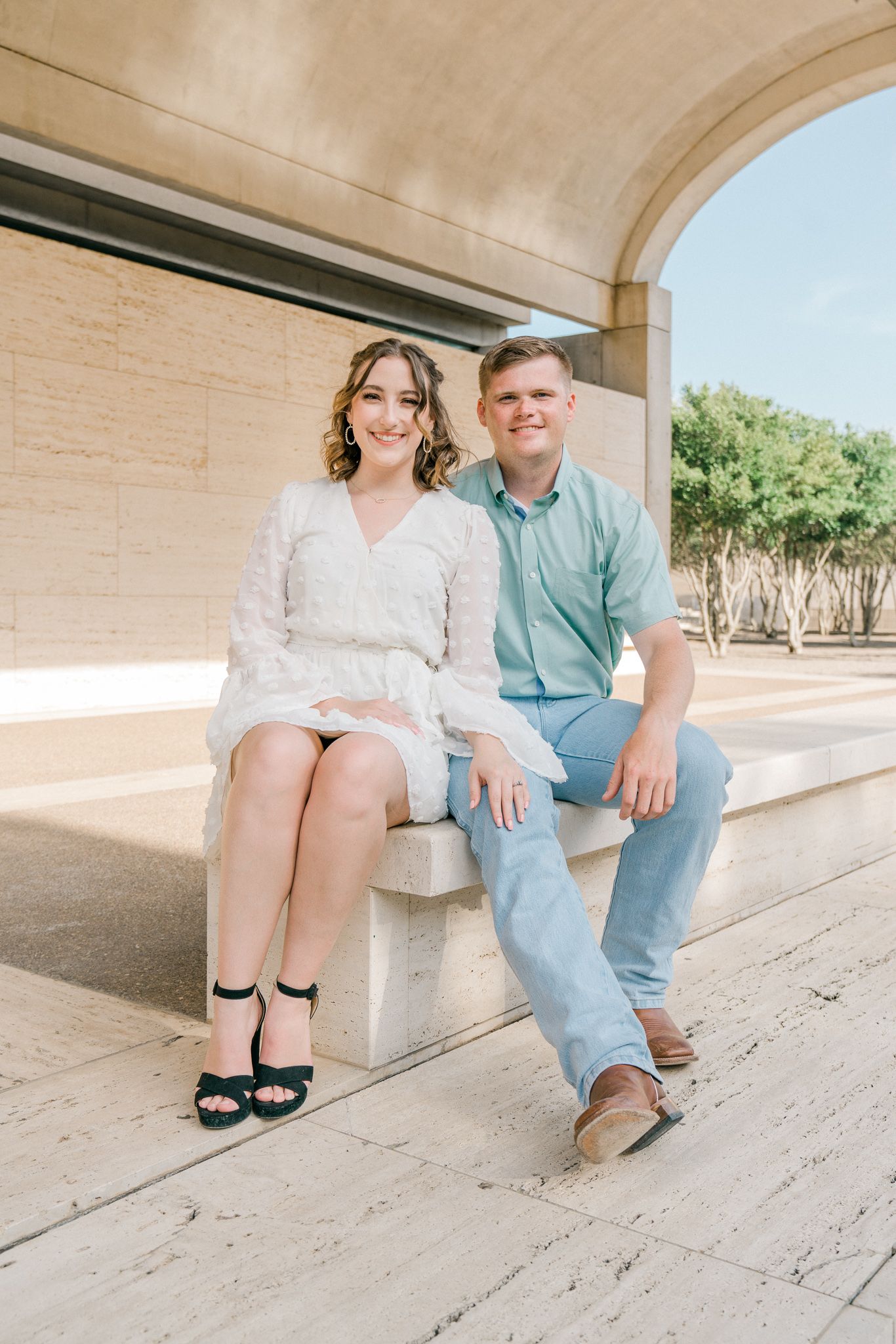 A couple sits together on a white stone bench, with their legs and feet in focus against an architectural backdrop.