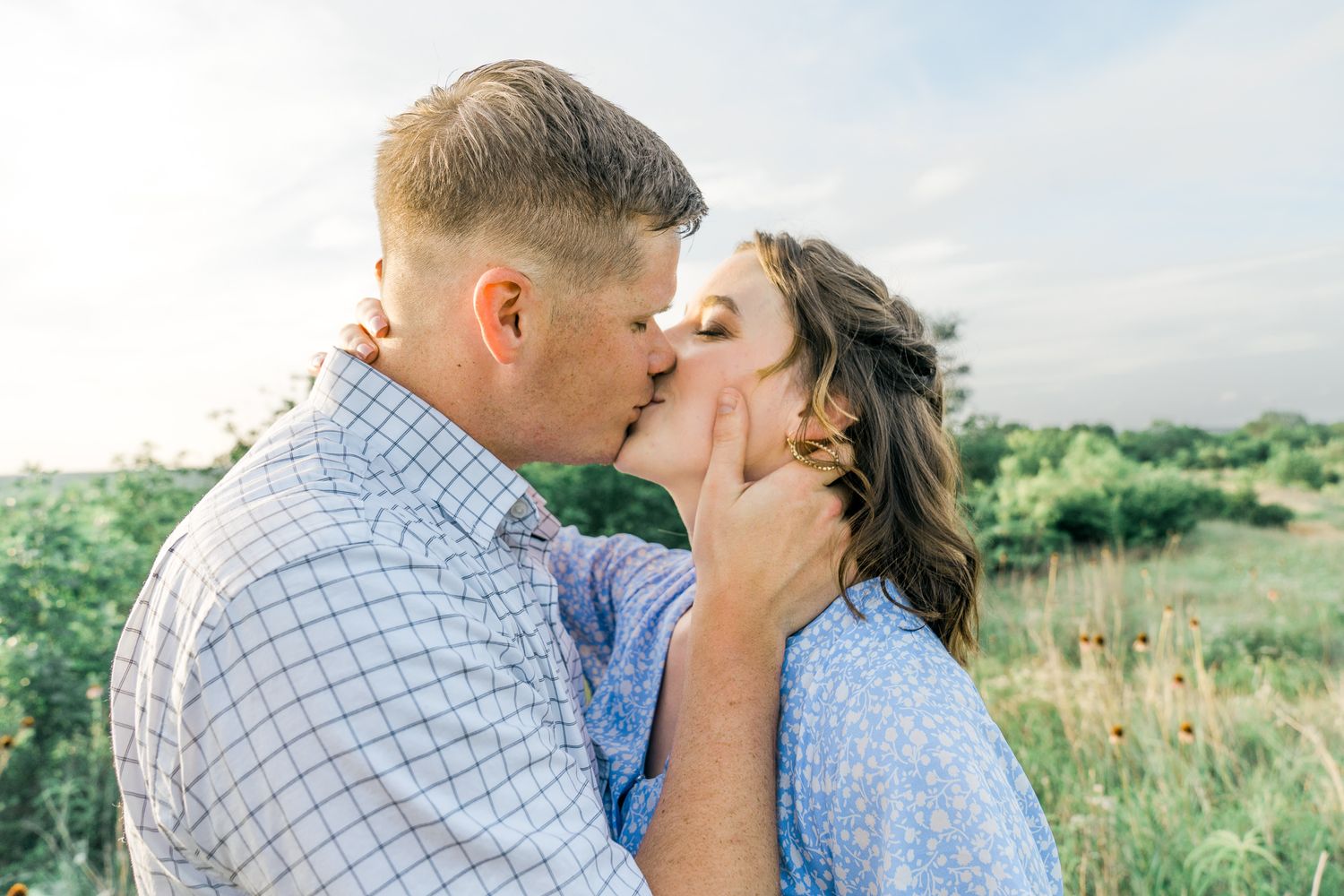 A romantic couple embraces and kisses against a bright blue sky in an outdoor setting.