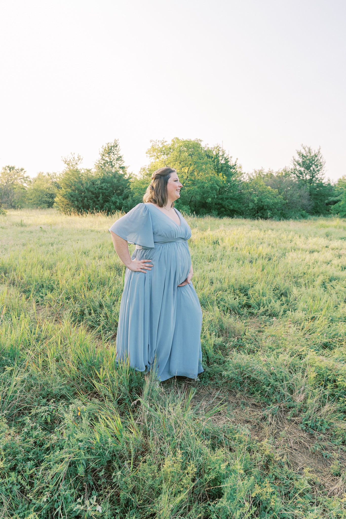 Maternity photo in golden field shows woman in light blue dress against sunset sky.
