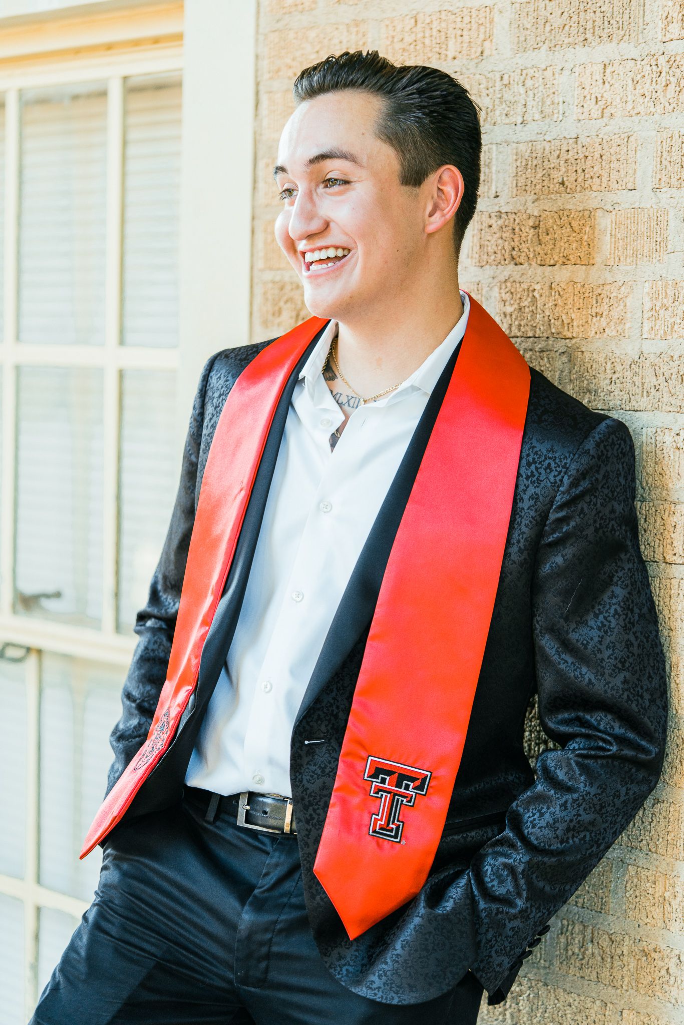 Graduate wearing black suit and bright red graduation stole leans against brick wall with a cheerful expression.