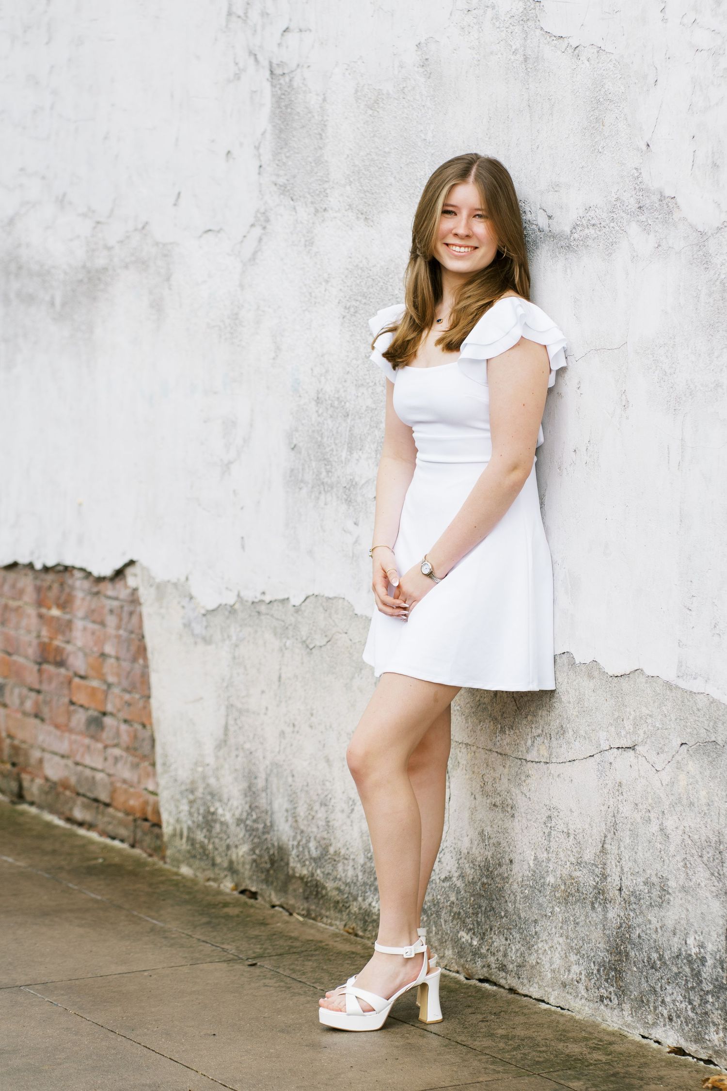 Young woman in white summer dress and platform sandals poses against textured wall with exposed brick.