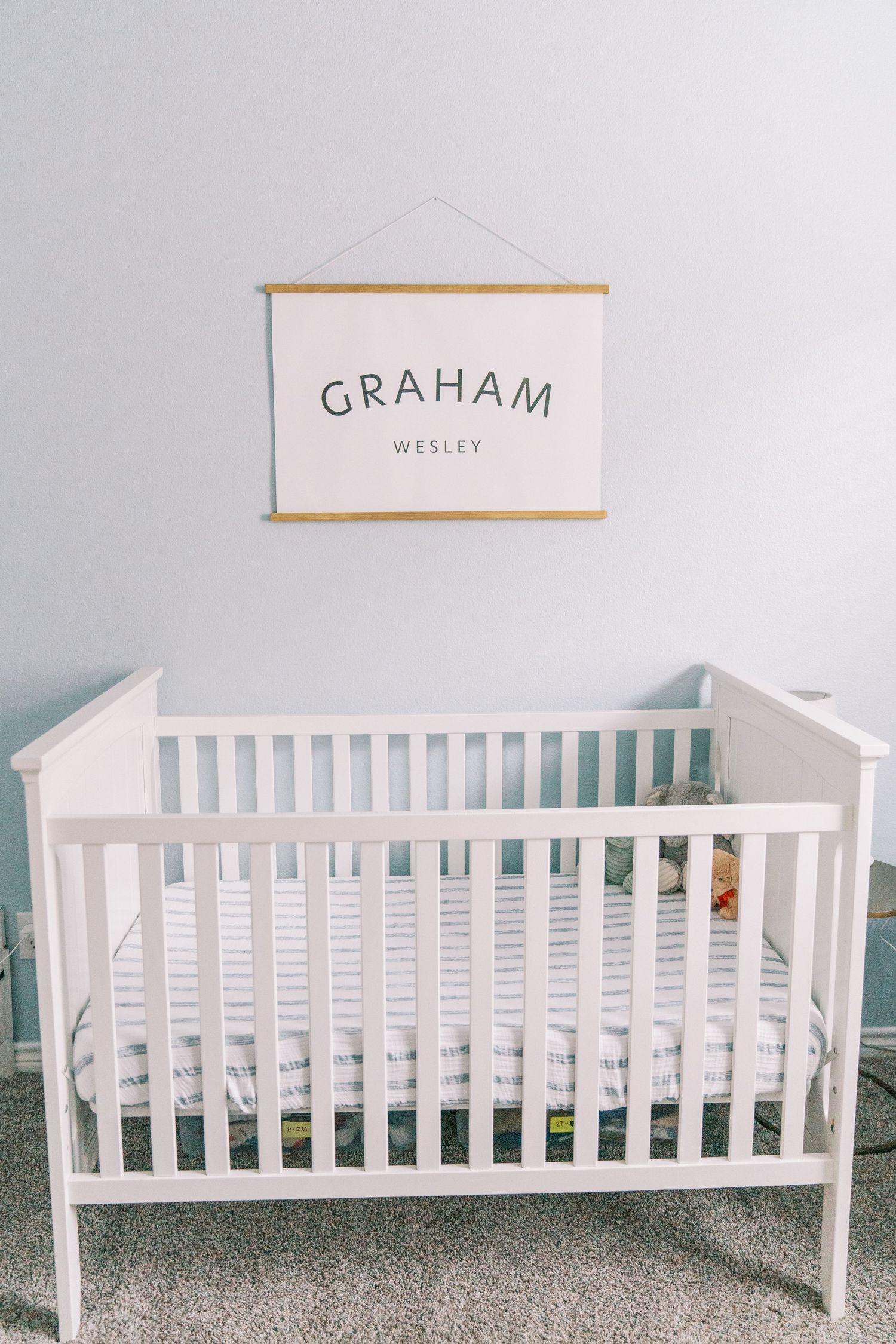 White wooden baby crib with name 'Graham Wesley' displayed on wall art above in minimalist nursery setting.