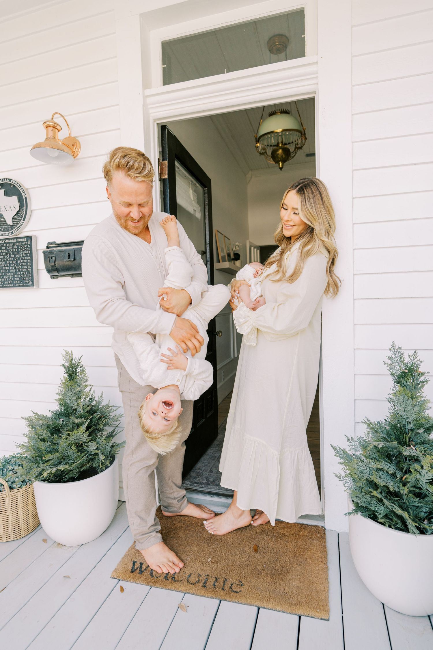 Family in white outfits sharing playful moments on a white porch with potted evergreens.