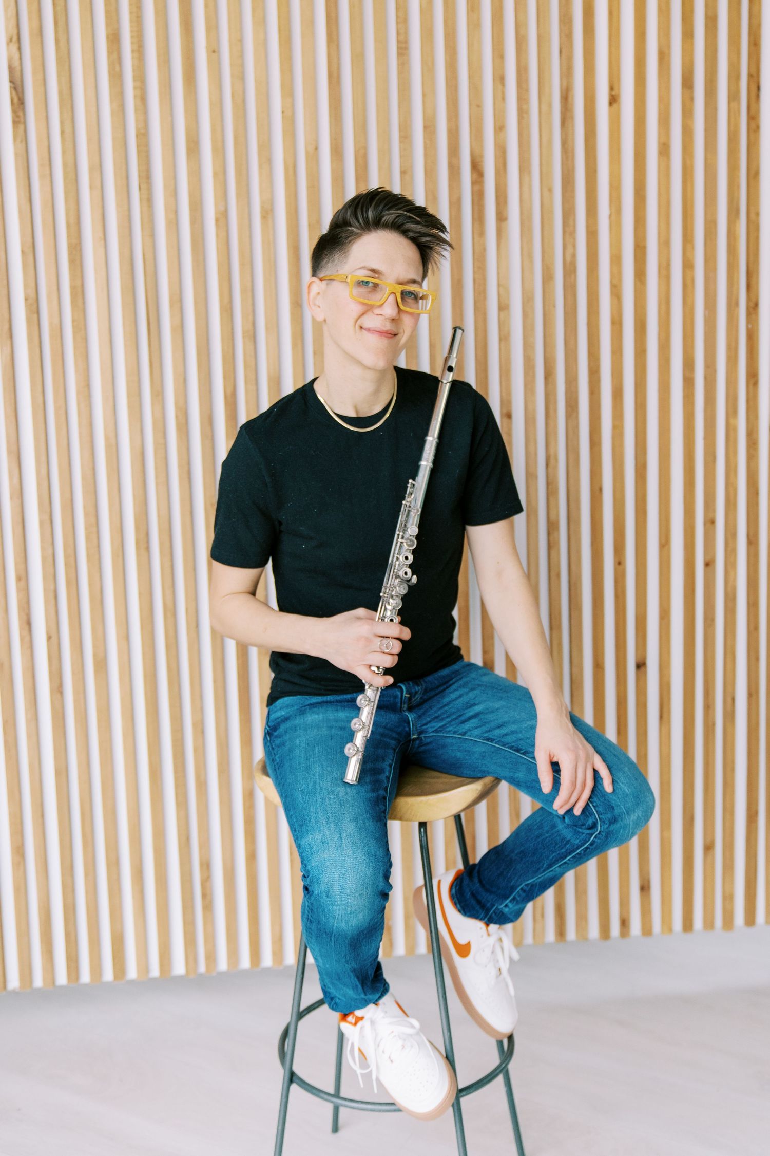Musician in black shirt and jeans sits on chair with flute instrument against striped wall background.