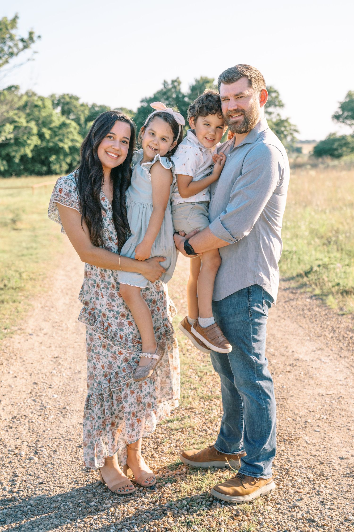 Happy family of four posing on a sunny dirt path surrounded by green fields and trees.