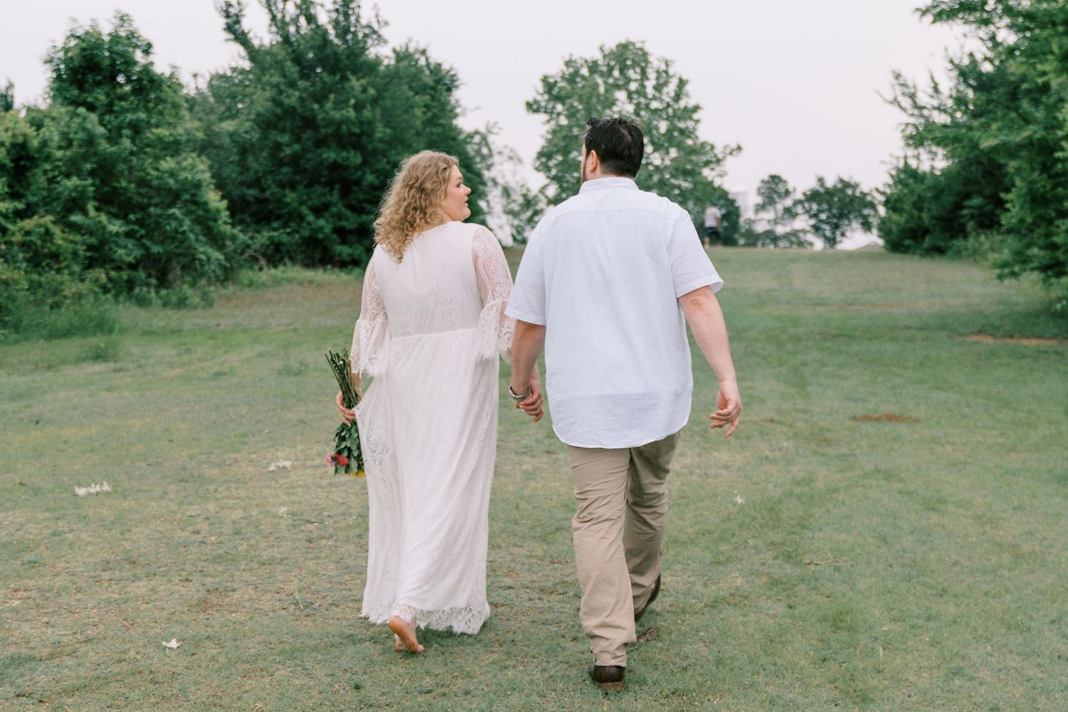 An engaged couple walks hand-in-hand through a green field, viewed from behind during a photo session.