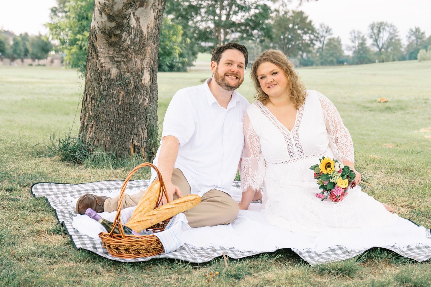 Couple in white attire sitting on picnic blanket under tree with wicker basket and sunflower bouquet.