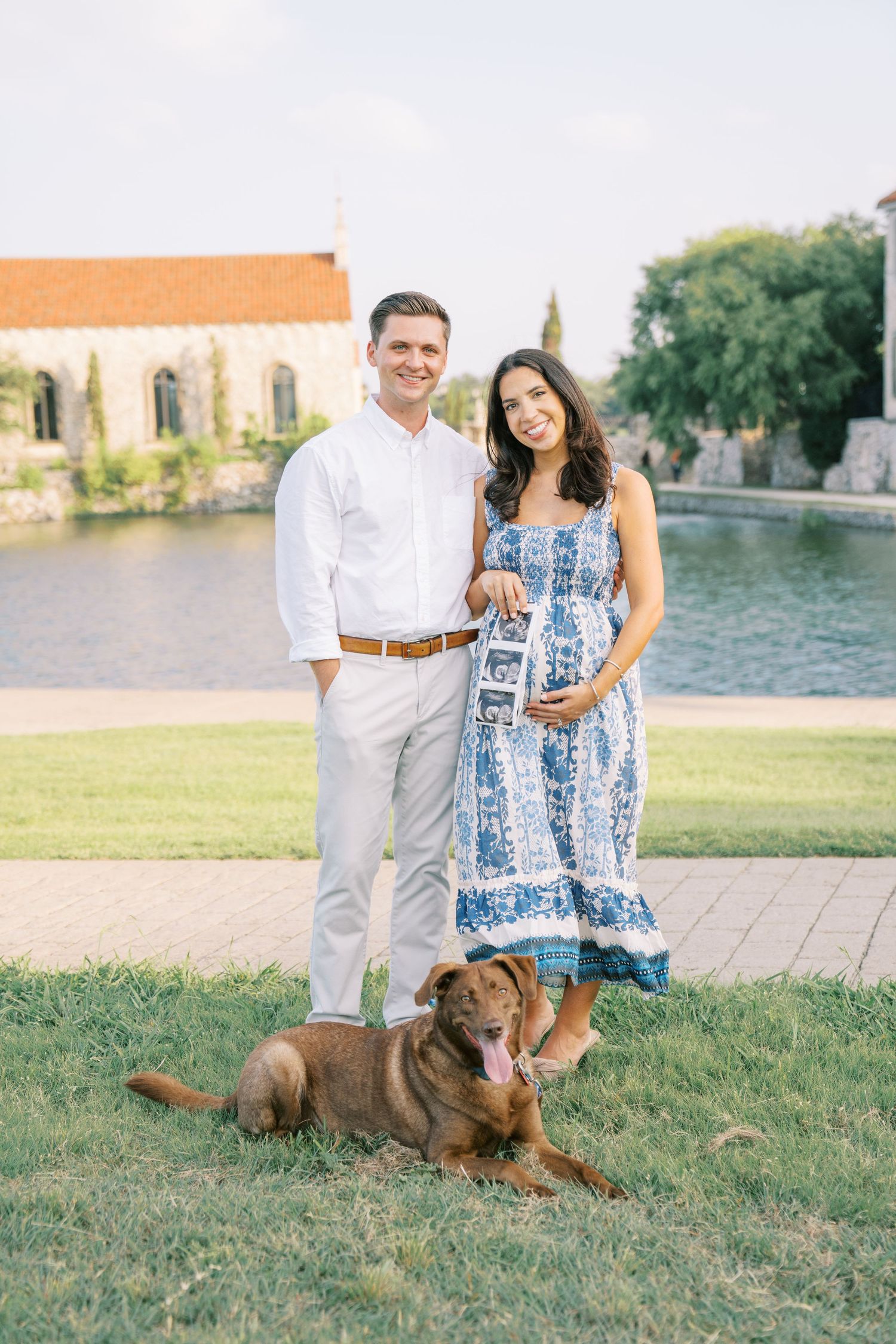 Couple holding ultrasound photo with brown dog on grass near pond, woman in blue floral dress, man in white shirt.