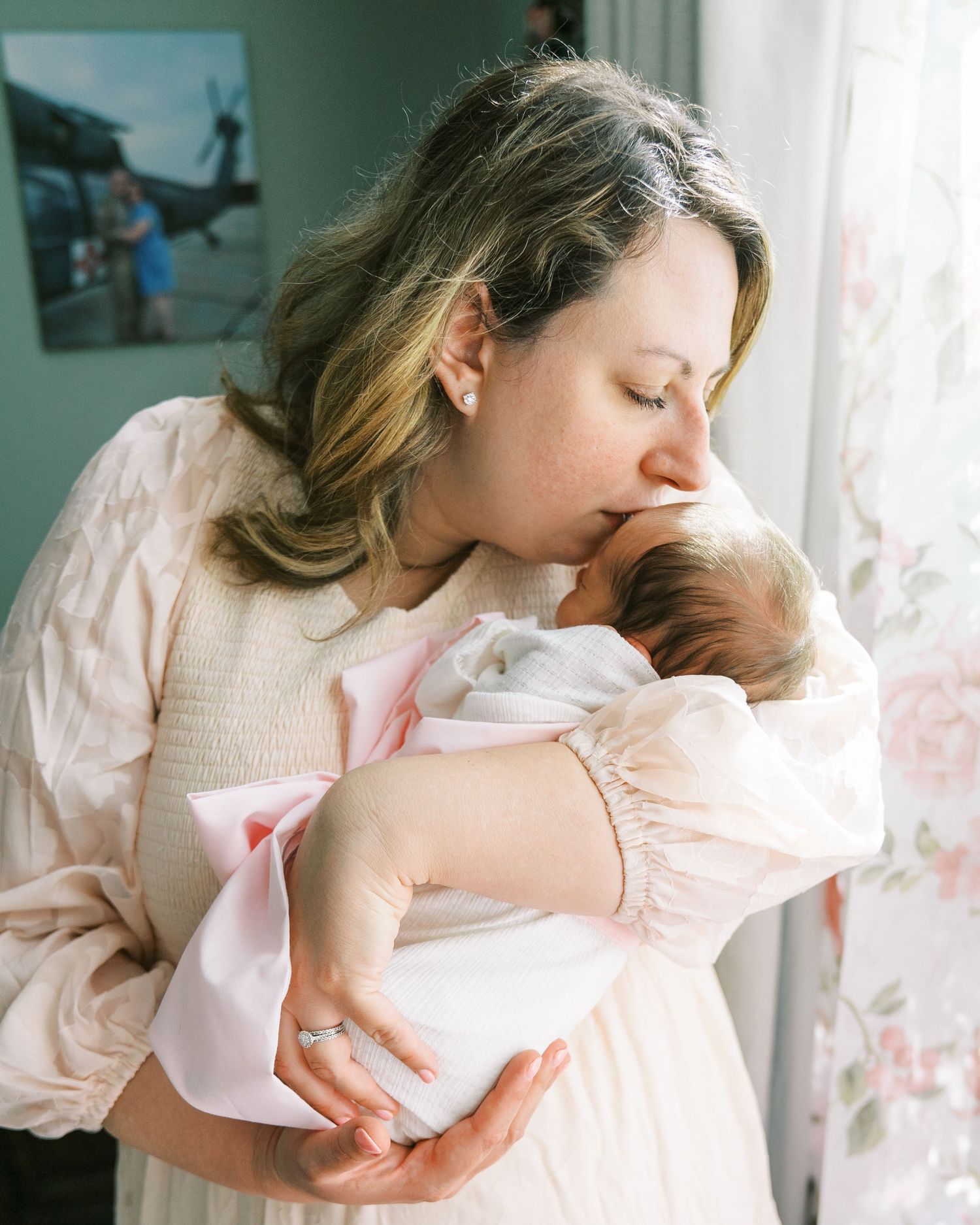 Gentle nurturing scene with a baby wrapped in pink near a window with floral curtains.