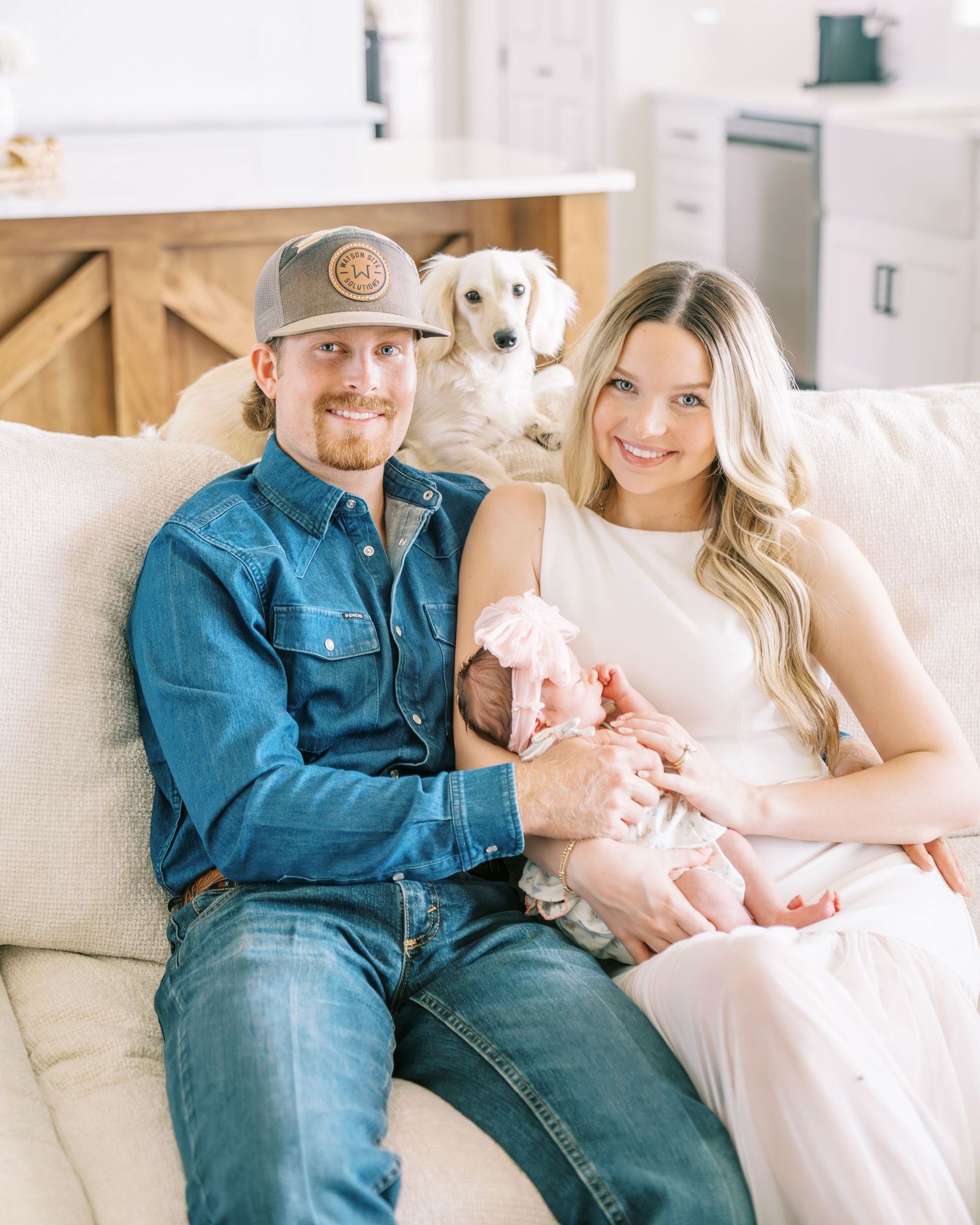 A young couple sits on a white couch holding their newborn baby, with a fluffy white dog peeking behind them.