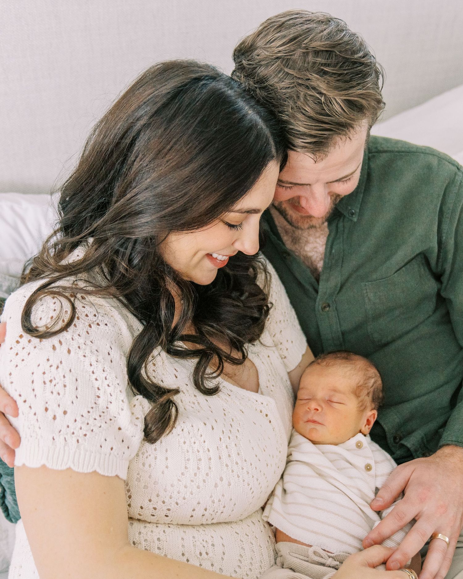 A couple in white and green clothing tenderly gazes at their sleeping newborn baby in a heartwarming family portrait.