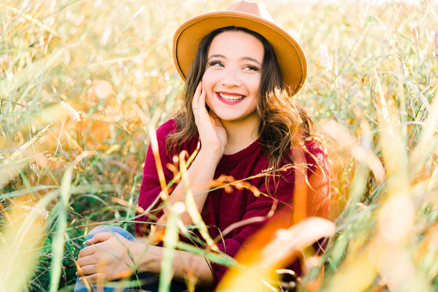 A person in a burgundy sweater and brown hat poses in a golden wheat field at sunset.