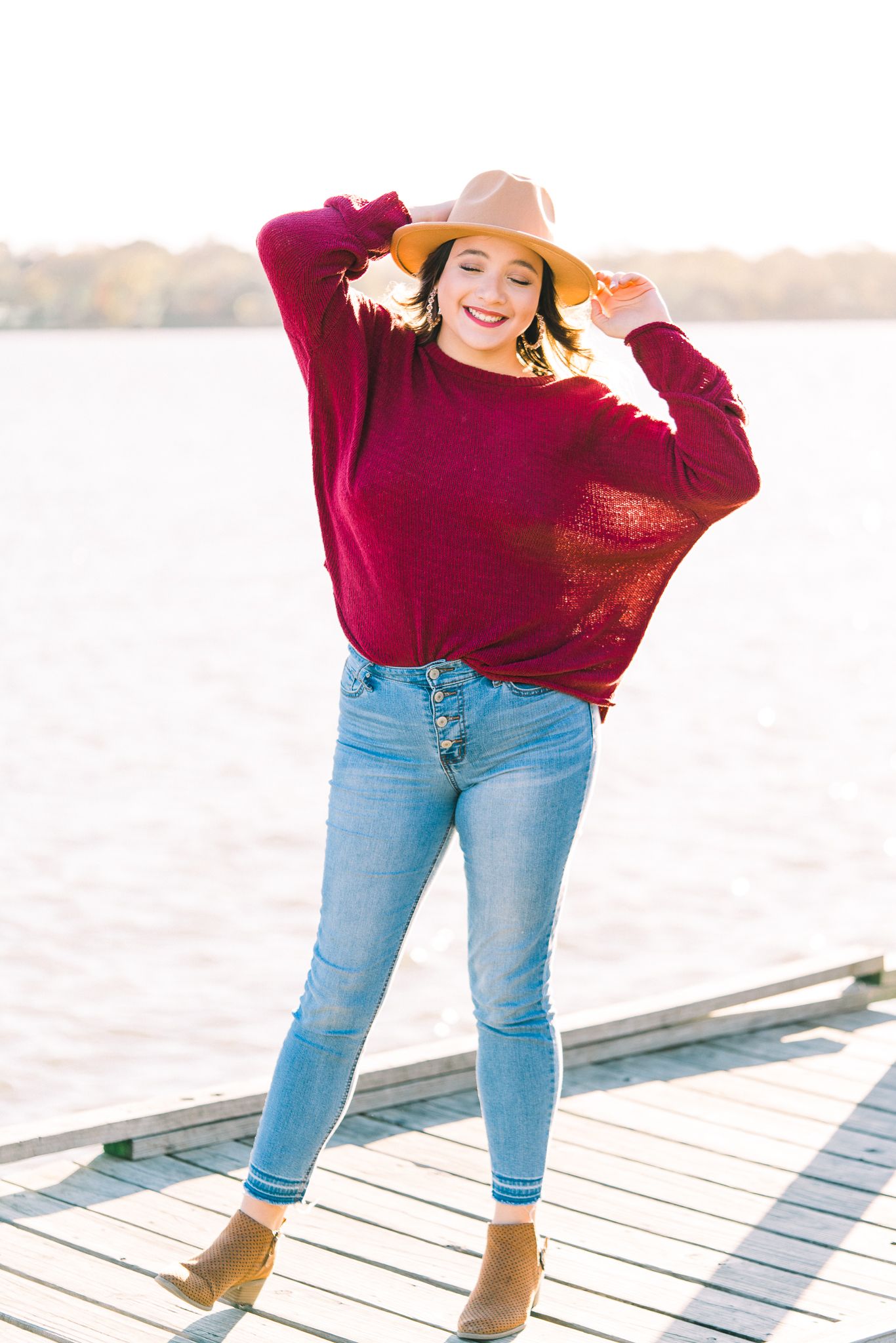 Person in burgundy sweater and jeans walks along wooden dock on sunny autumn day wearing tan wide-brim hat.