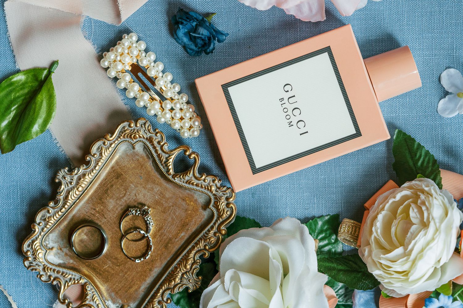 Wedding details flatlay with perfume bottle, rings on ornate tray, and white roses on blue background.
