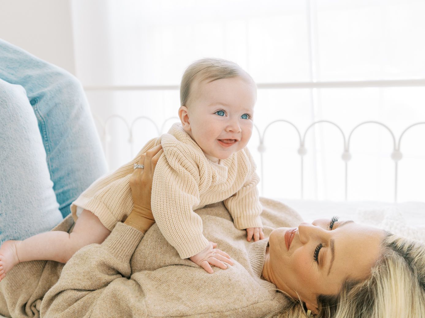 Mother and baby in coordinating beige sweaters sharing joyful moments on a white bed.