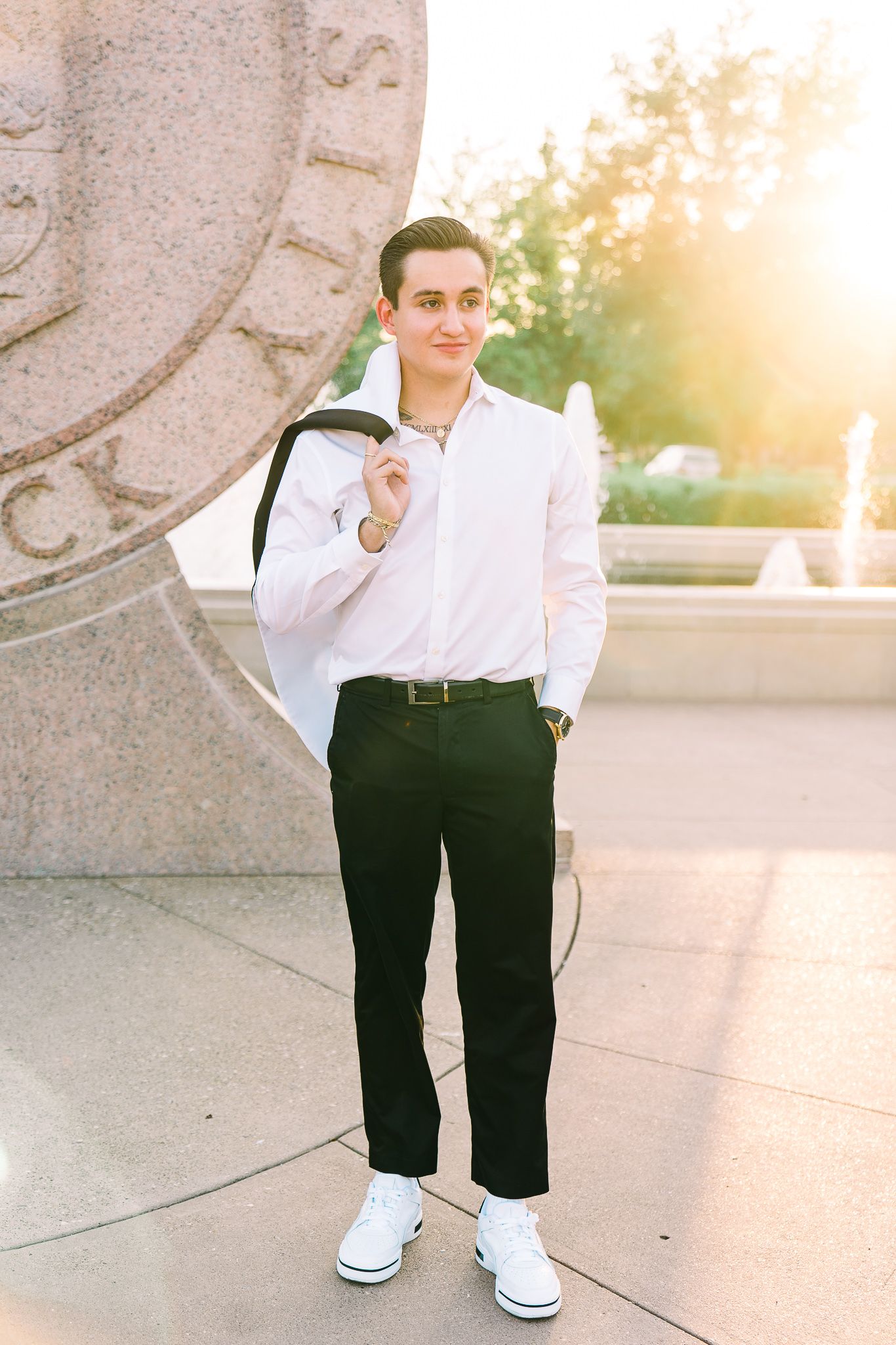A person in a white dress shirt and black pants poses casually against a stone wall in warm sunset lighting.