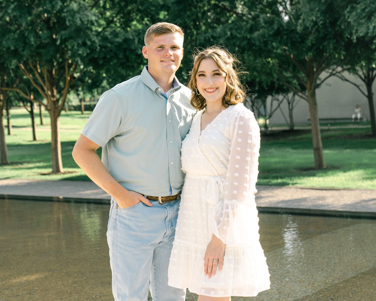 A couple poses together outdoors in coordinating light-colored outfits on a sunny day in a park setting.