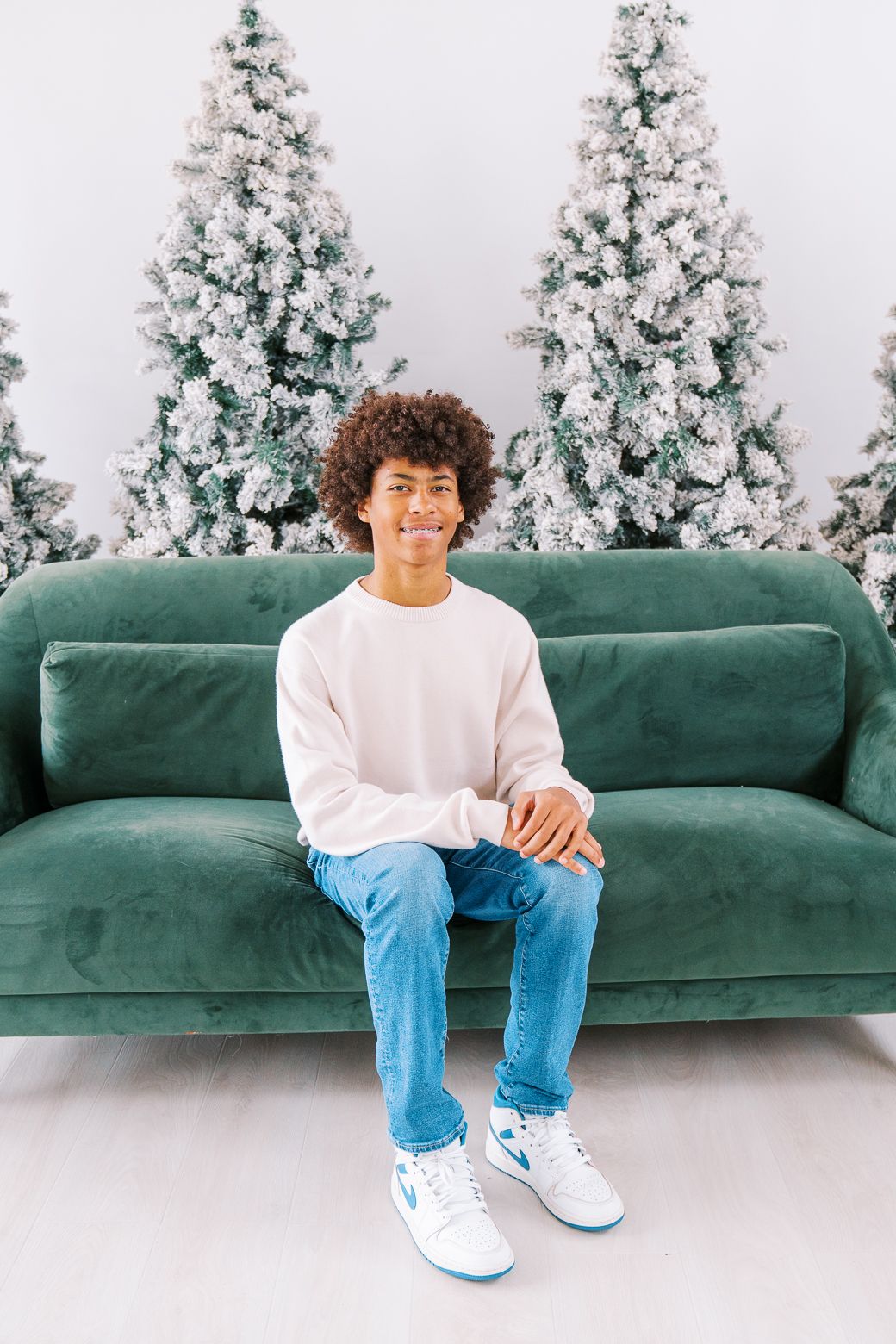 Person in white sweater and jeans sitting casually on green velvet couch with flocked Christmas trees behind.