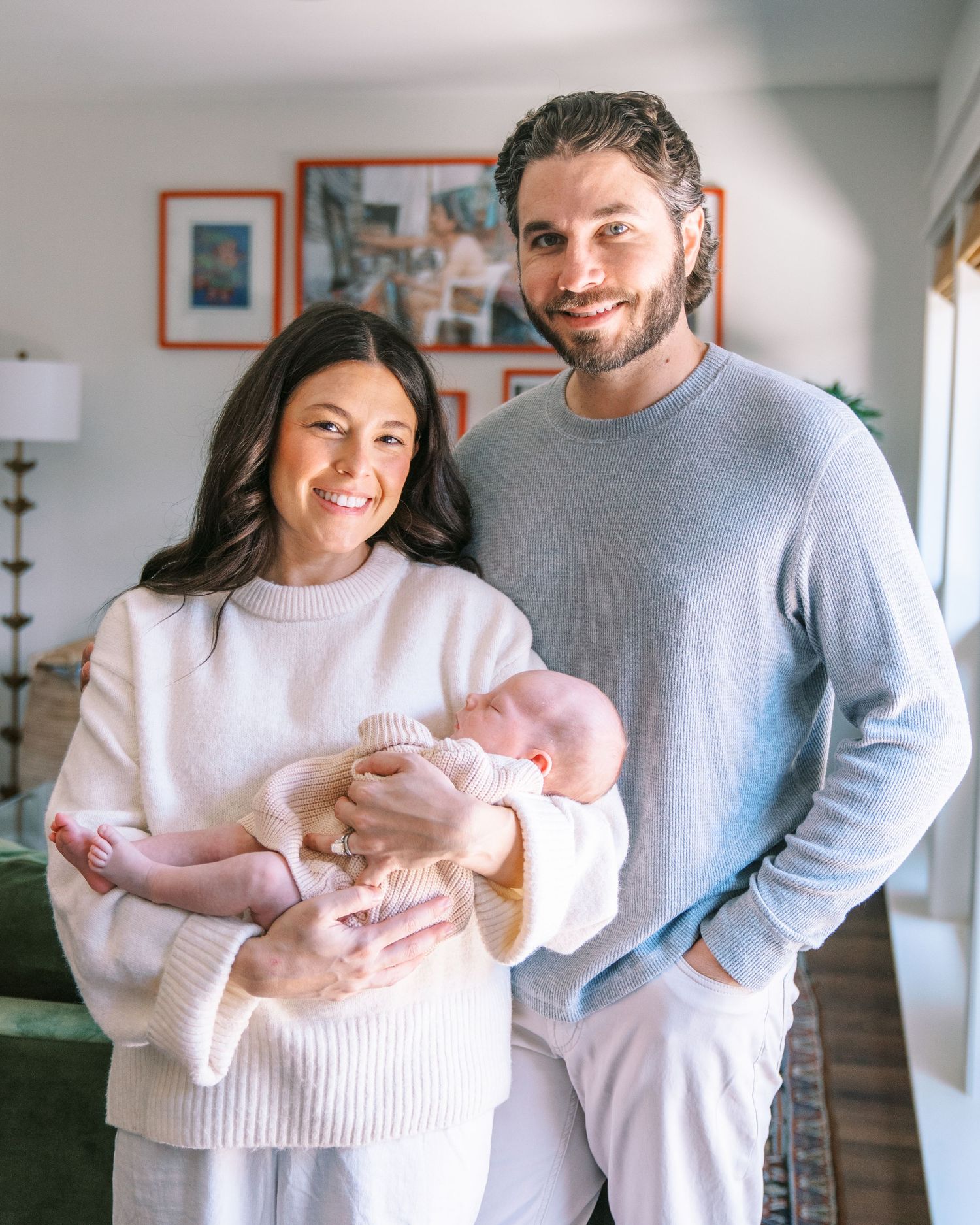 Smiling couple in cozy home holding their newborn baby, dressed in white and gray sweaters.