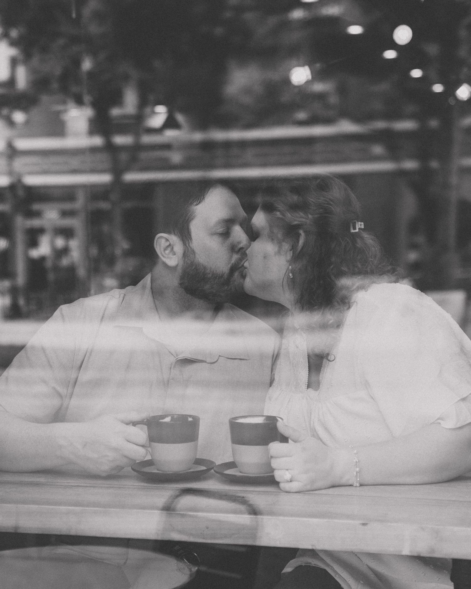 Black and white shot of couple kissing through cafe window with coffee cups on table.
