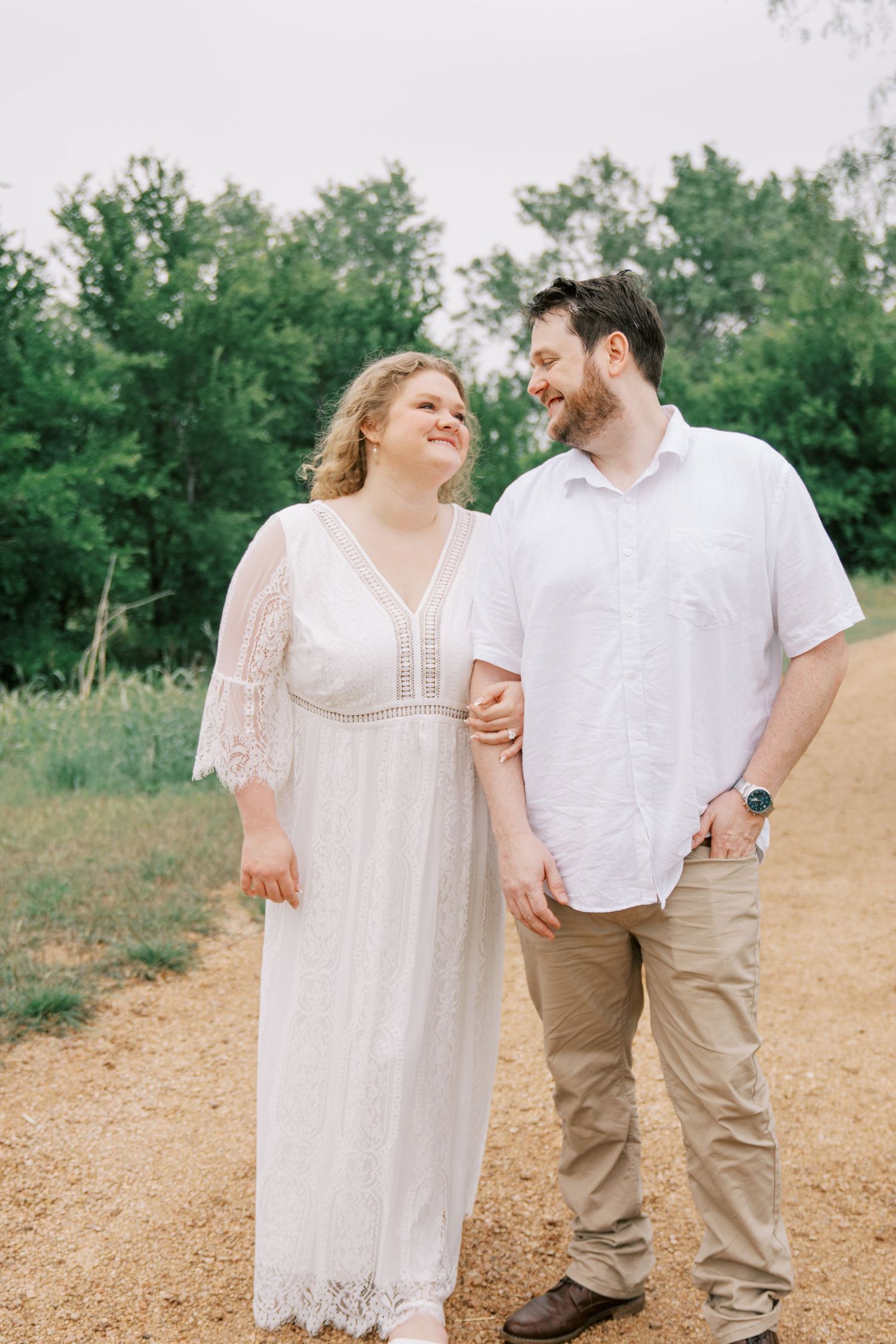 Engaged couple in white dress and shirt walking hand in hand on a dirt path surrounded by green trees.