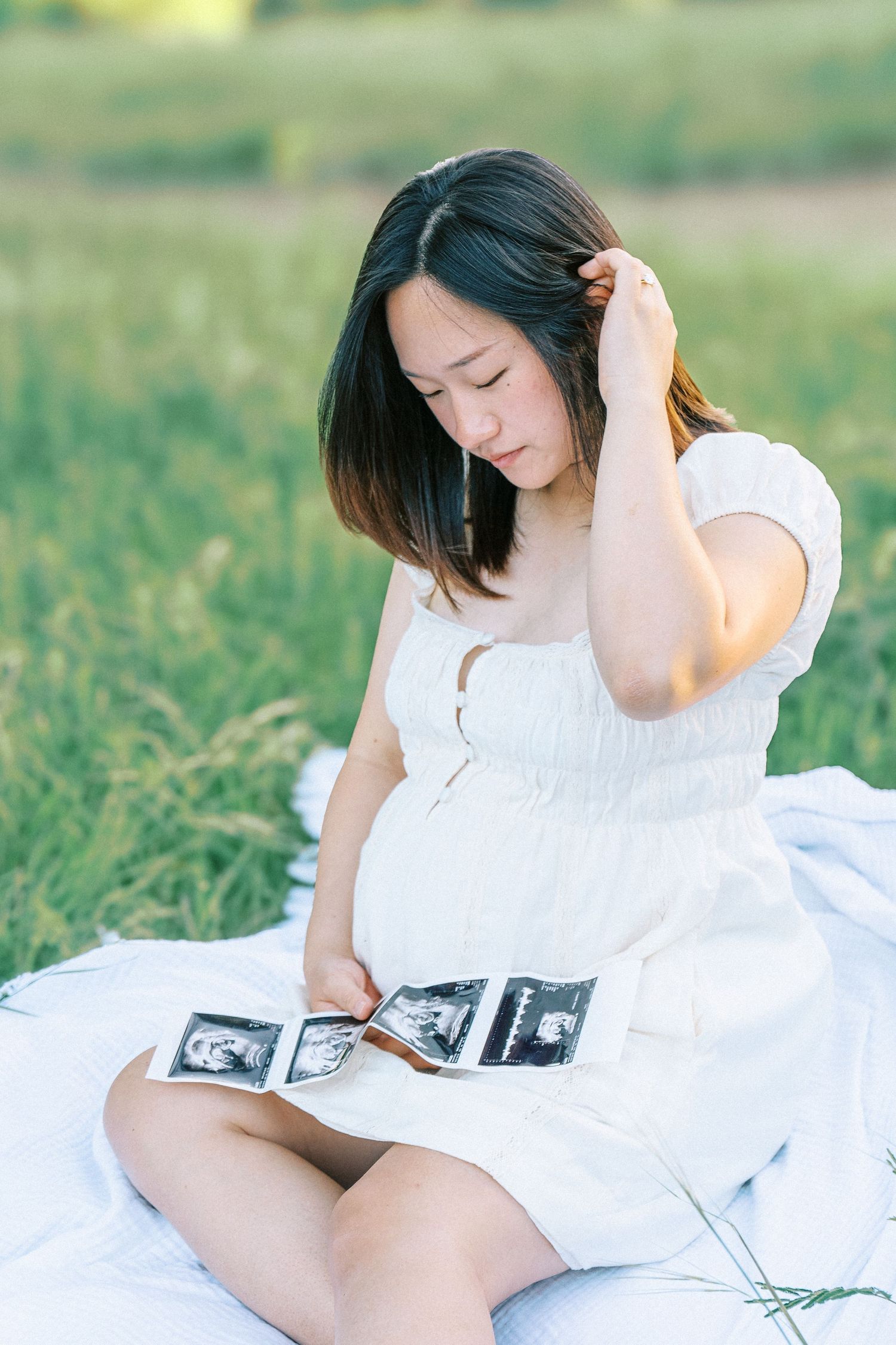 Pregnant Asian woman in white dress sitting on blanket in field, looking at ultrasound photos.