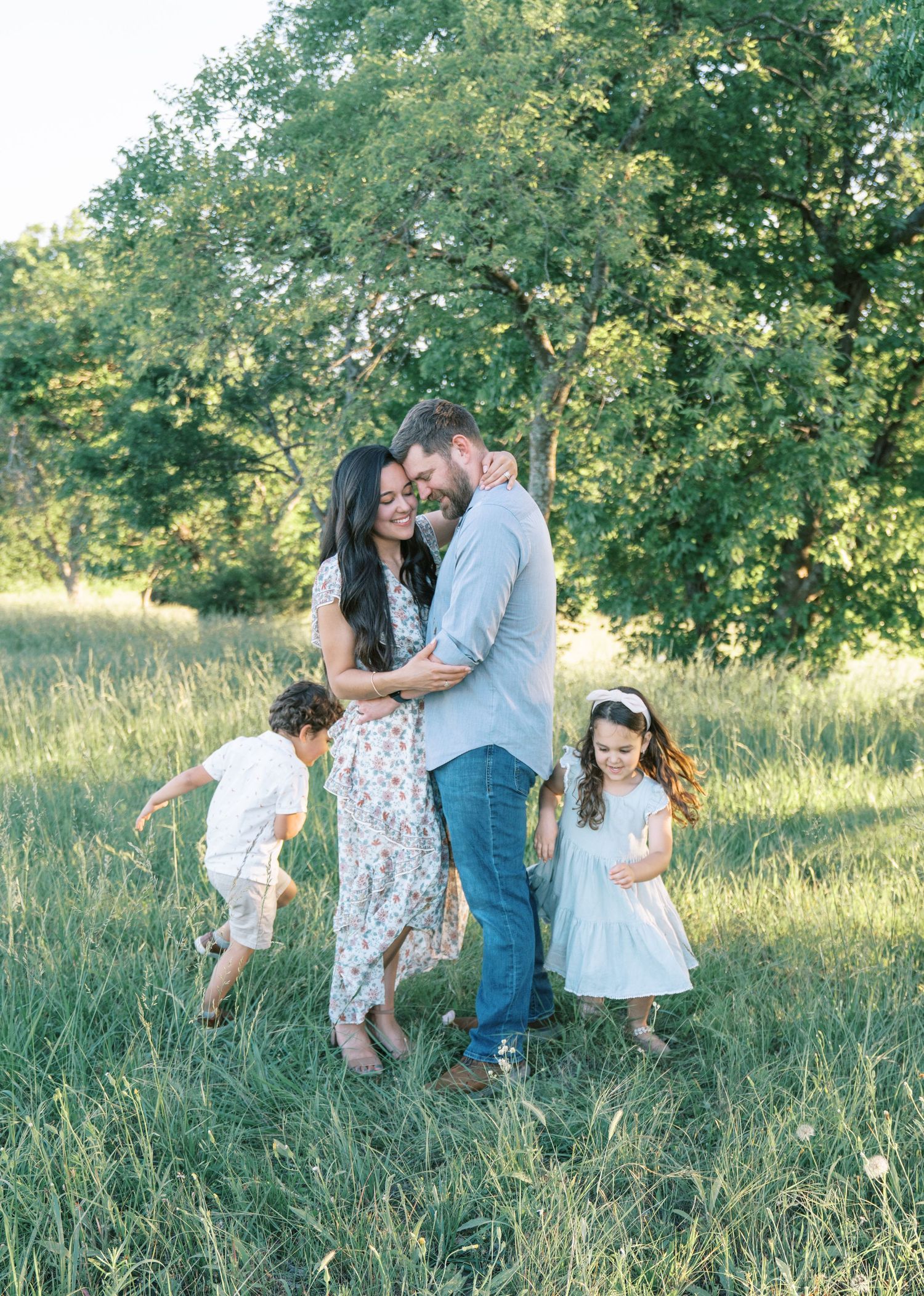 Family of four posing in a sunny green meadow, parents embracing while two young children play around them.