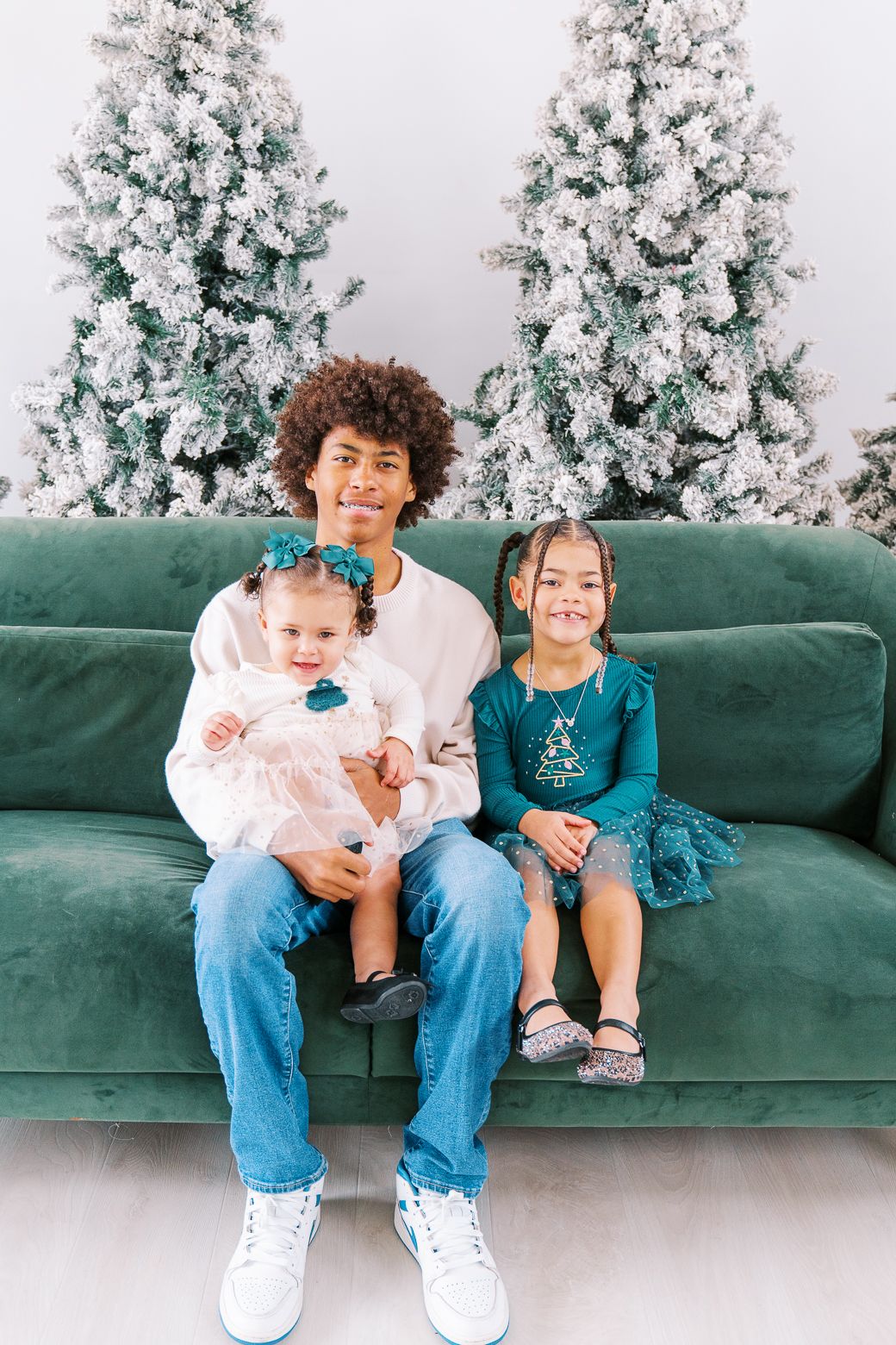 Young man sits on green velvet sofa holding two little girls in front of snow-flocked Christmas trees.