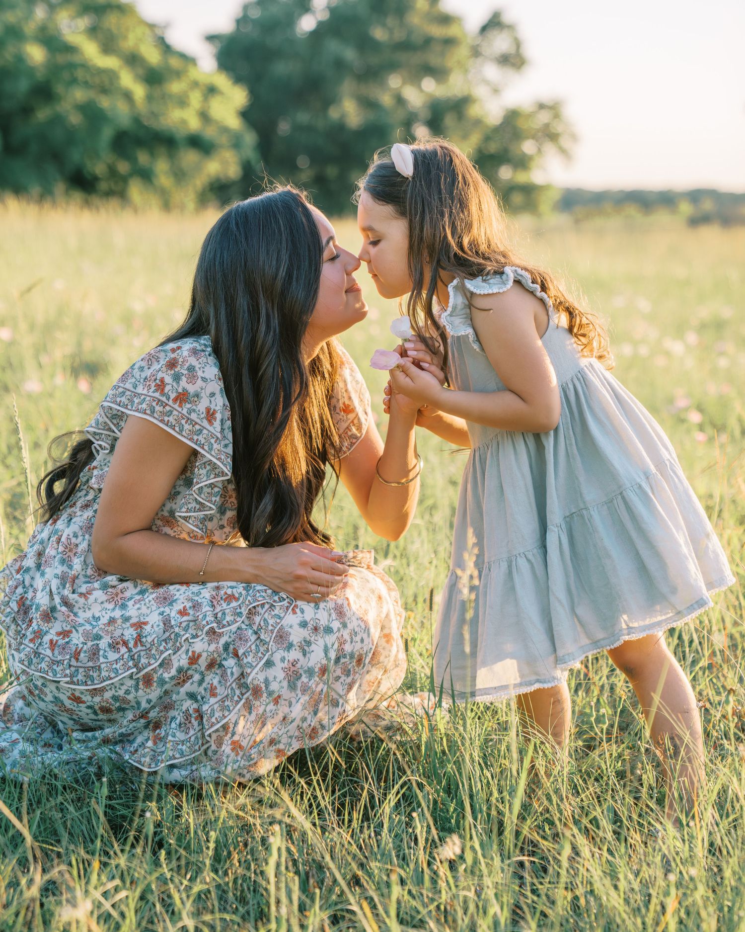 A mother and young daughter share a tender nose-to-nose moment in a sunlit meadow, both wearing floral and pastel dresses.