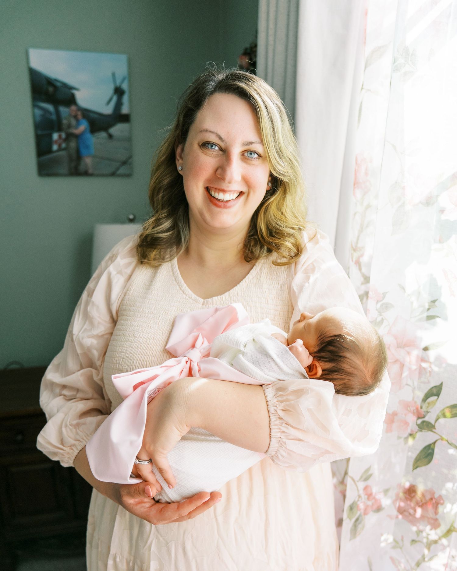 A tender moment in a nursery with sage green walls as a woman in pink dress holds a swaddled newborn.