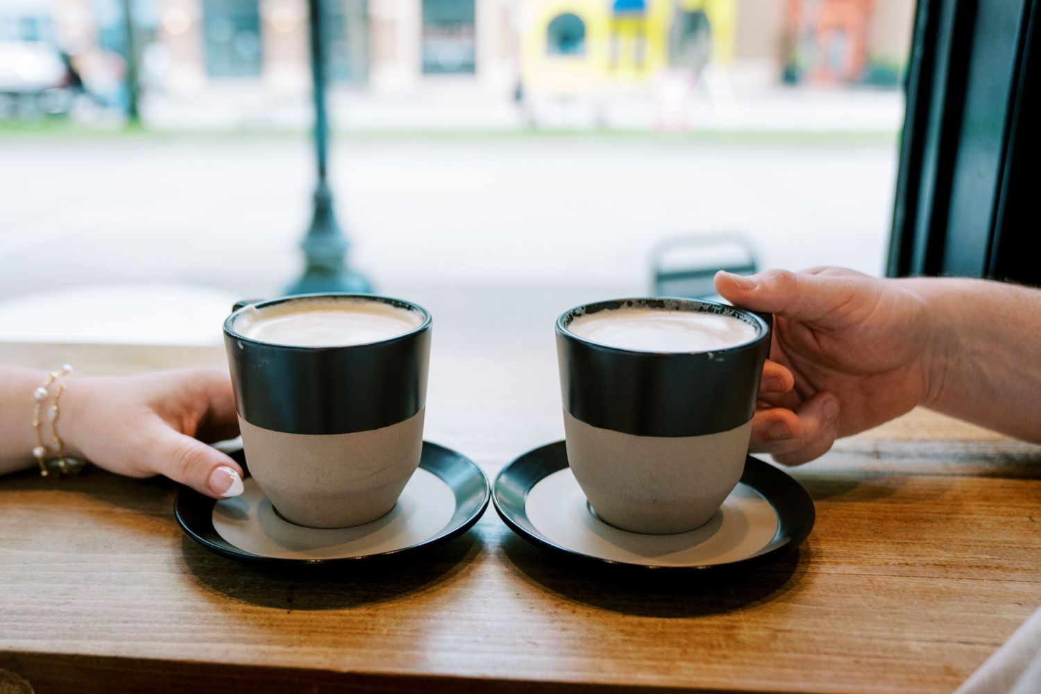 Two lattes in modern cups on saucers held by a couple at a wooden cafe table.