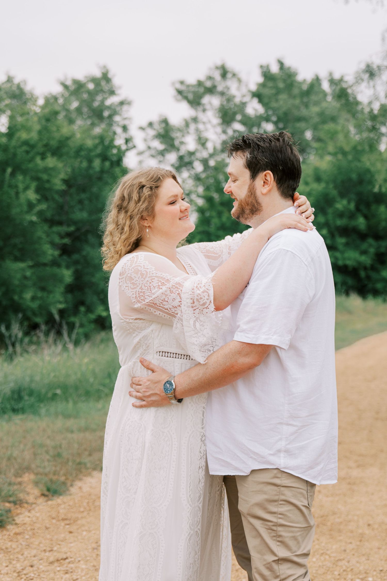 An engaged couple embraces on a dirt path surrounded by lush green trees during an outdoor photo session.