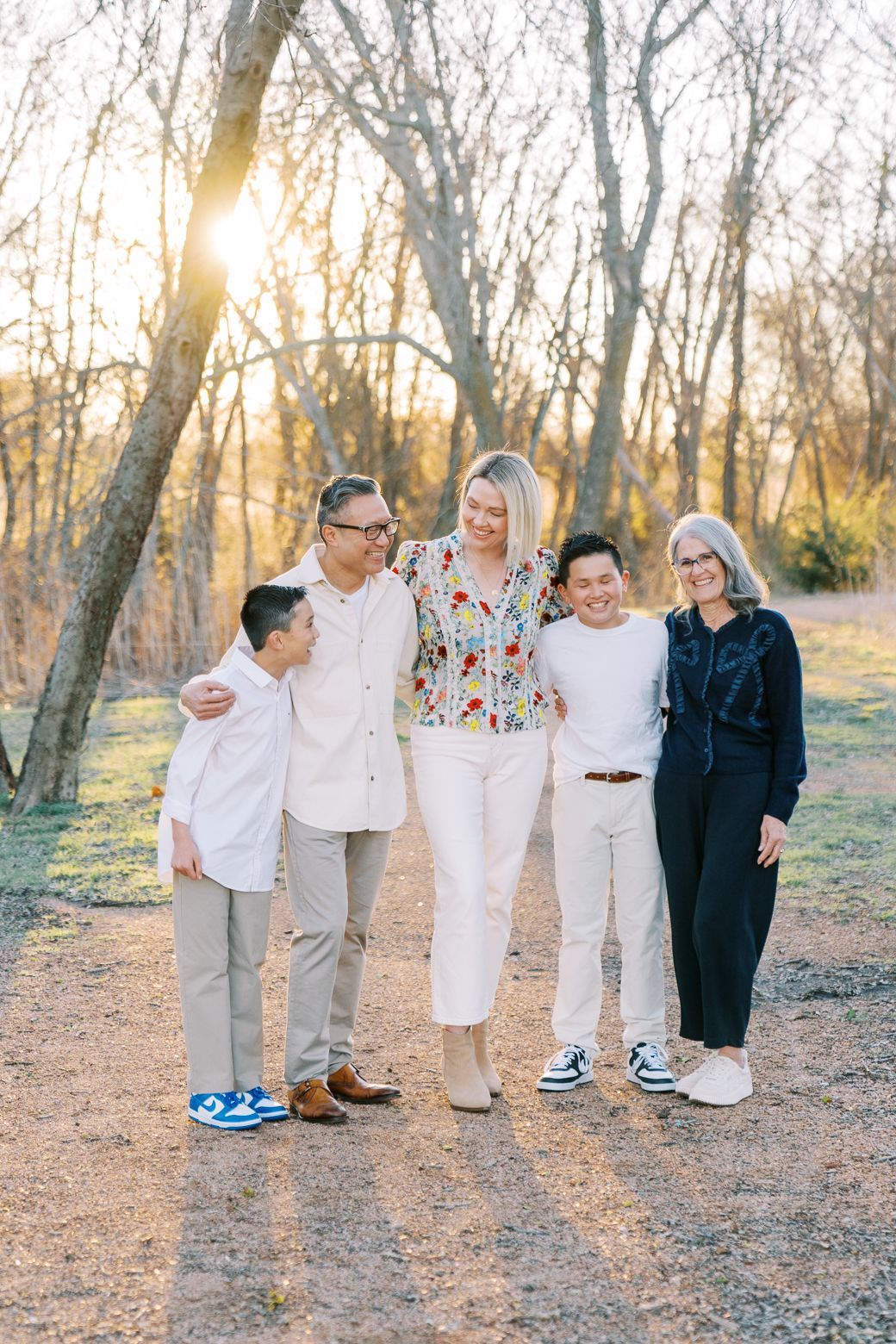 Family of five smiling together on a dirt path at golden hour, surrounded by bare trees in a park setting.