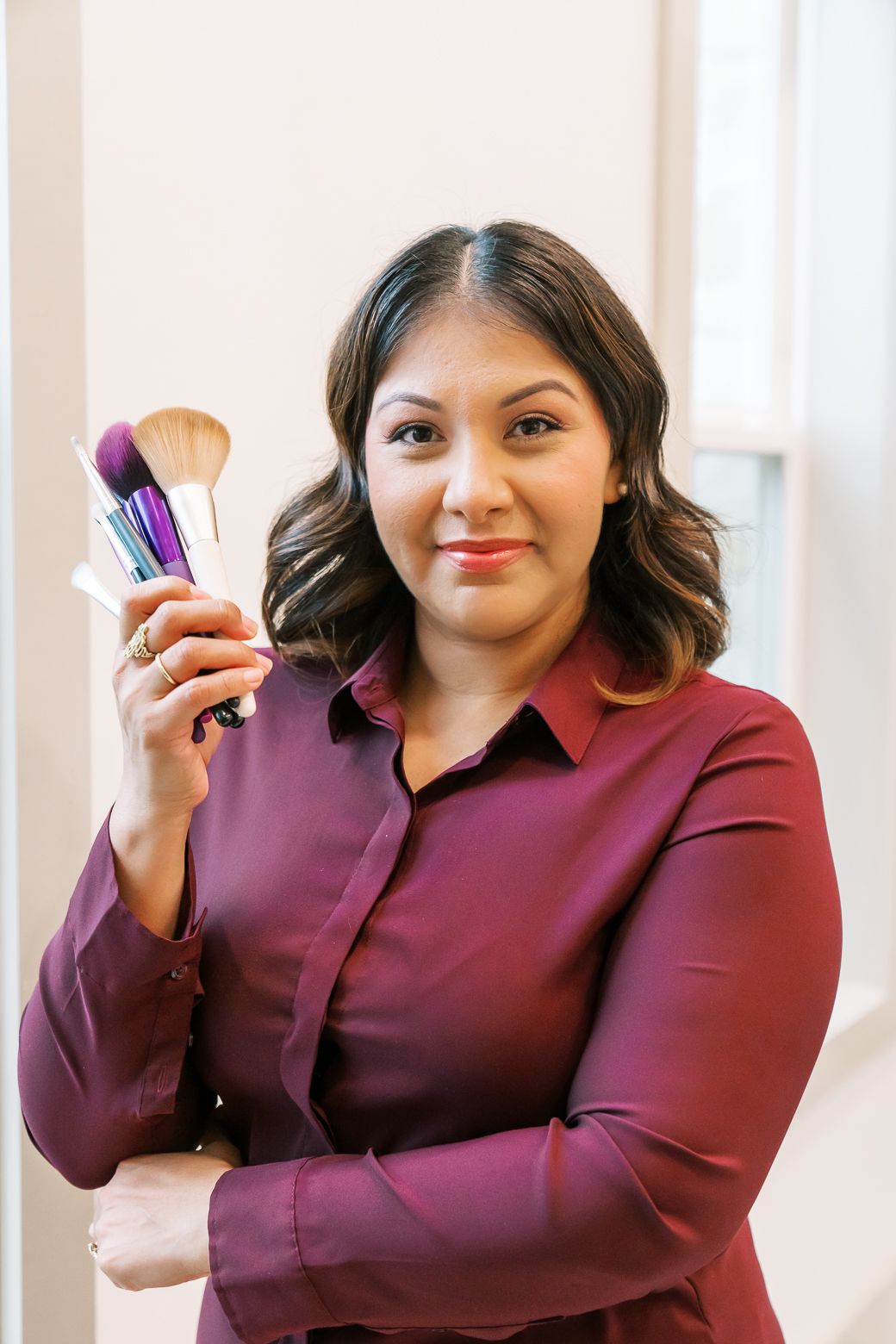 A person in a burgundy blouse holds makeup brushes while posing in a bright interior setting.