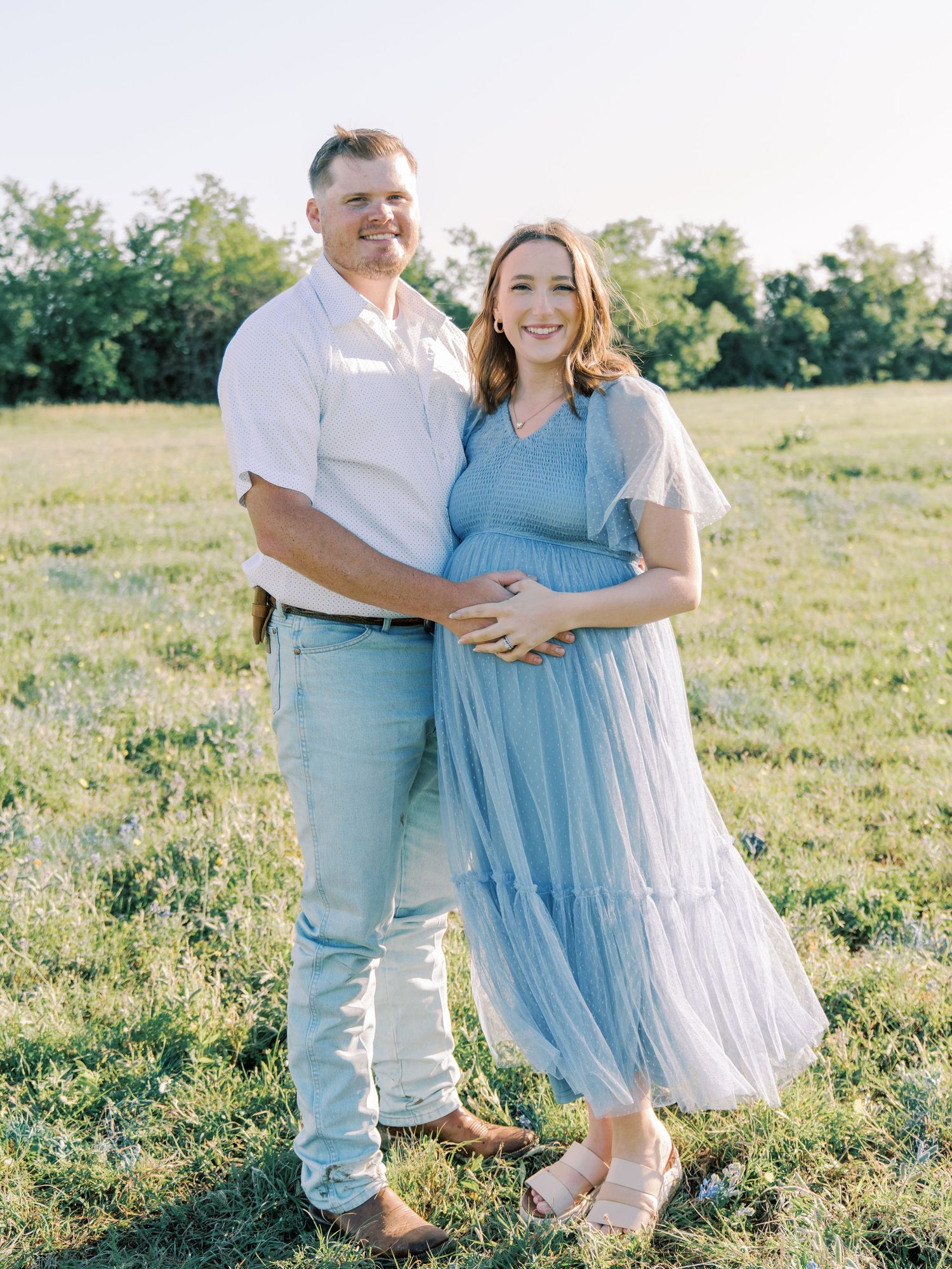 Expectant couple embraces in a sunlit field, wearing coordinating blue and white outfits for maternity photos.
