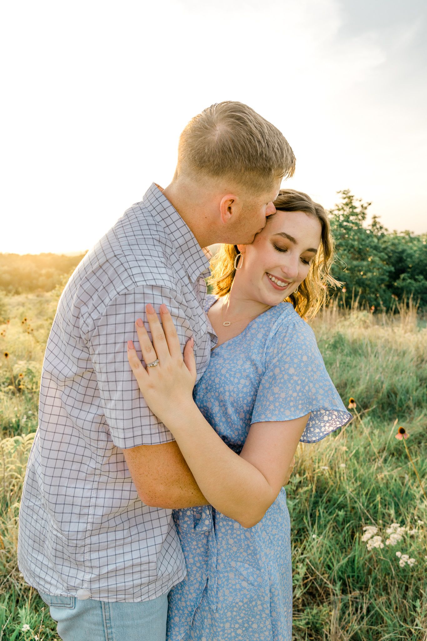 A romantic couple embraces in a golden field during sunset while showing off an engagement ring.