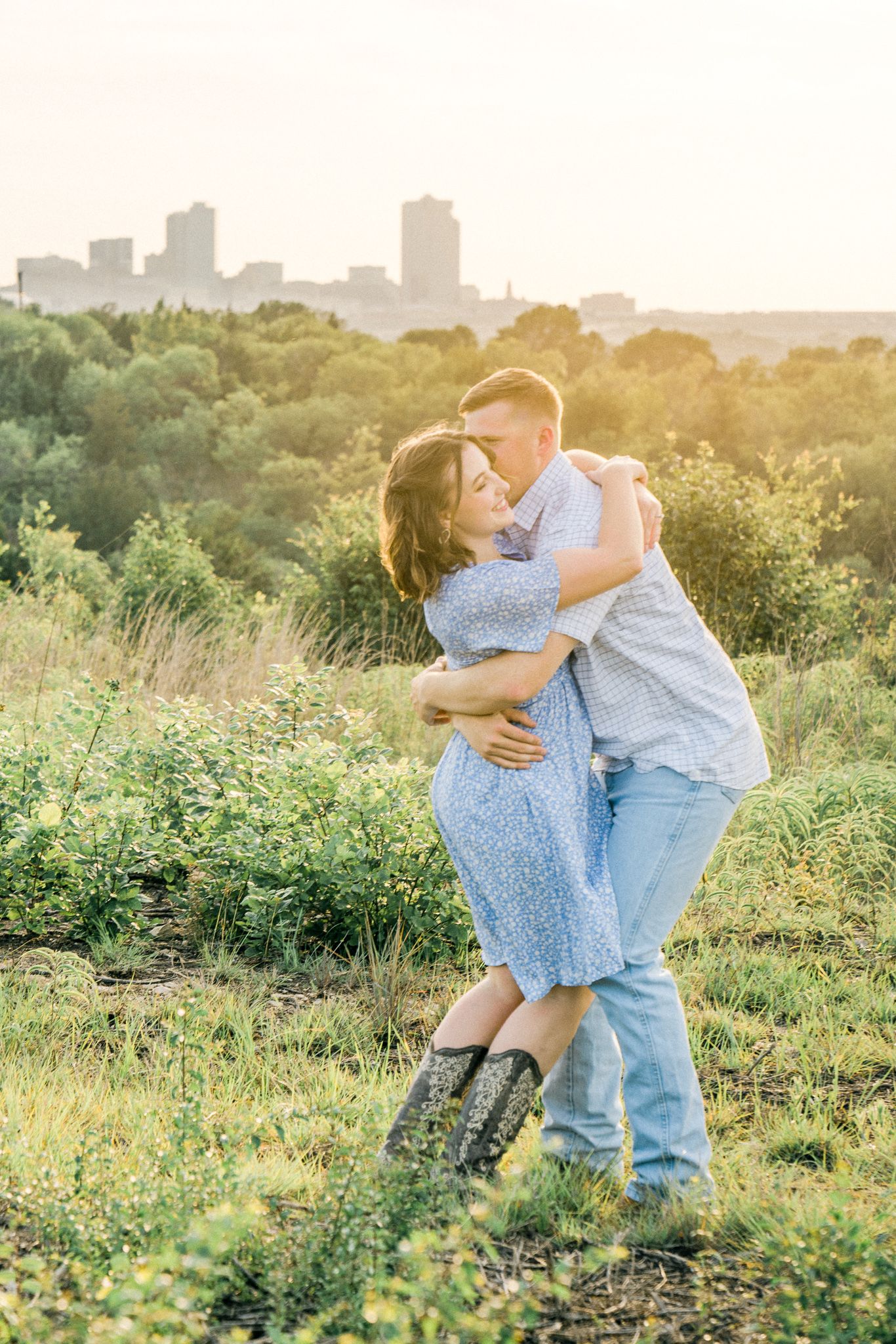 Couple in coordinating blue outfits embrace in a sunlit meadow with wildflowers and city skyline in background.