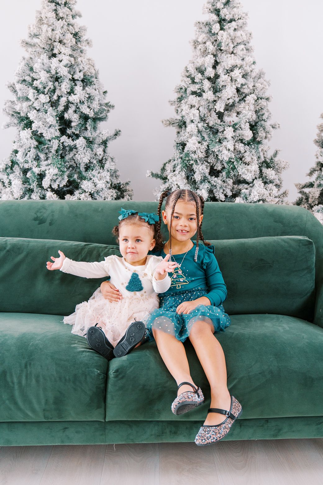 Two children sit playfully on a green velvet couch with snow-covered Christmas trees in the background.