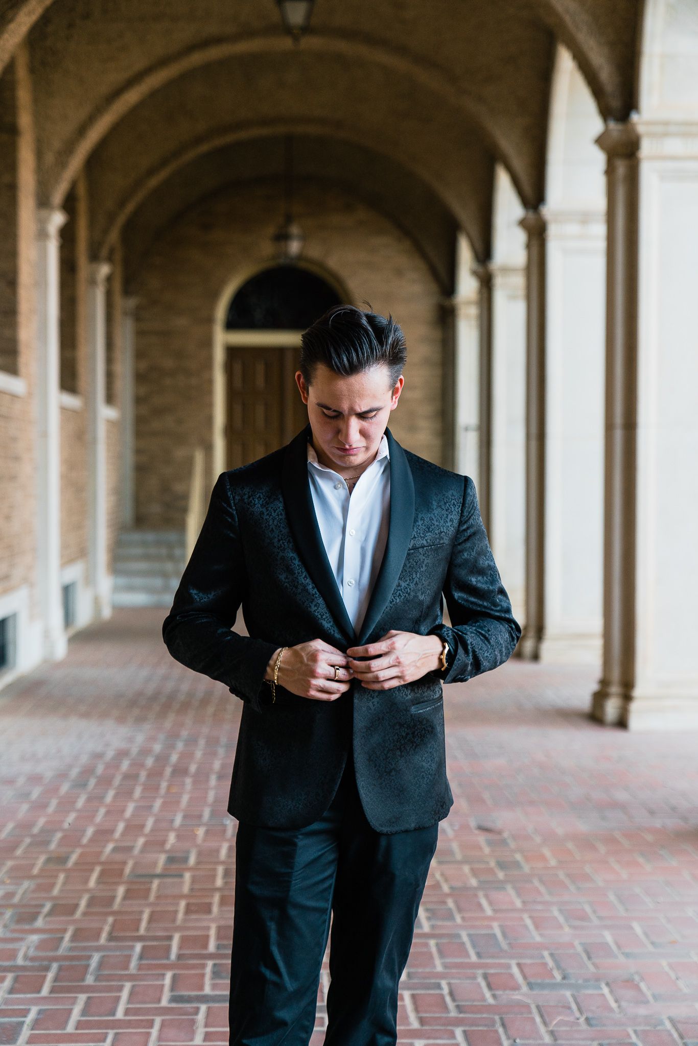 Professional in dark suit checks phone while walking through elegant arched colonnade hallway.