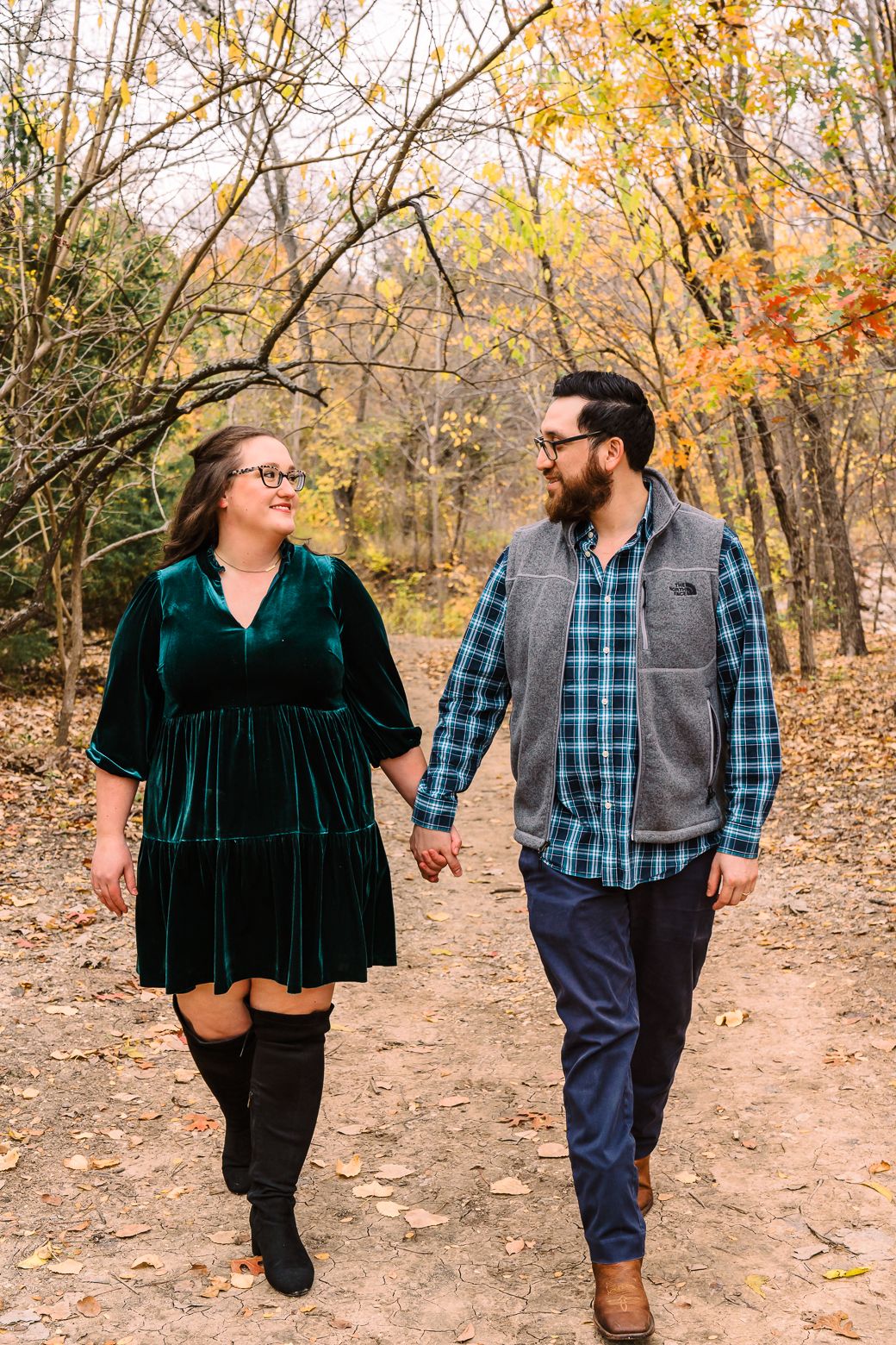 A couple walks hand in hand on a forest trail surrounded by golden autumn foliage and fallen leaves.