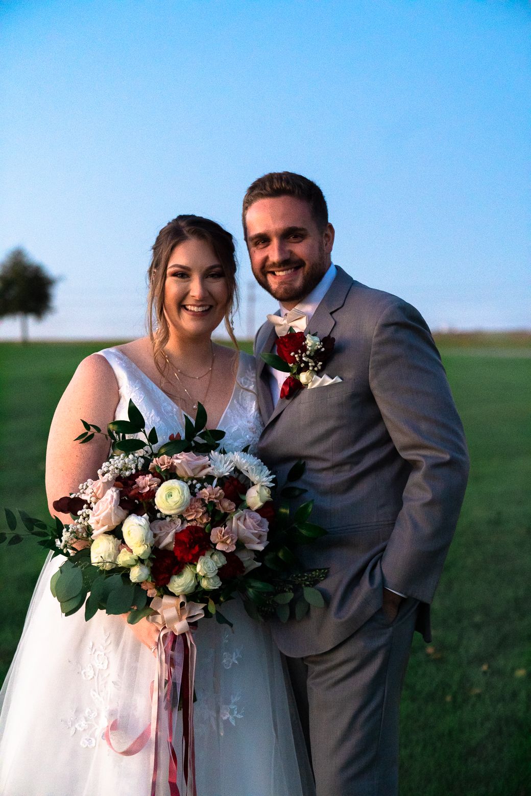 A couple in wedding attire embraces in a grassy field under blue sky with beautiful floral bouquet.