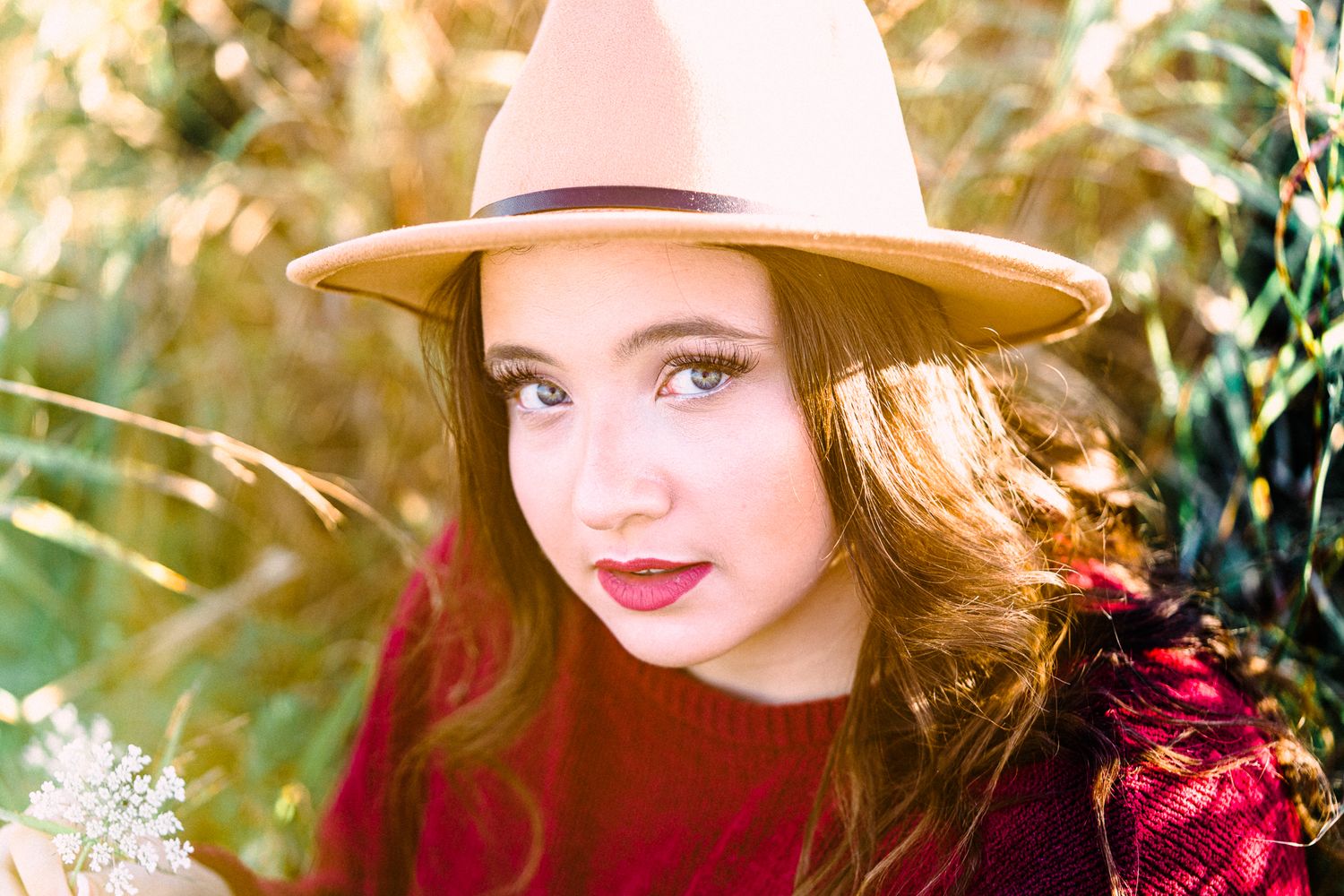 A person in a burgundy sweater and beige hat poses outdoors among sunlit autumn foliage.