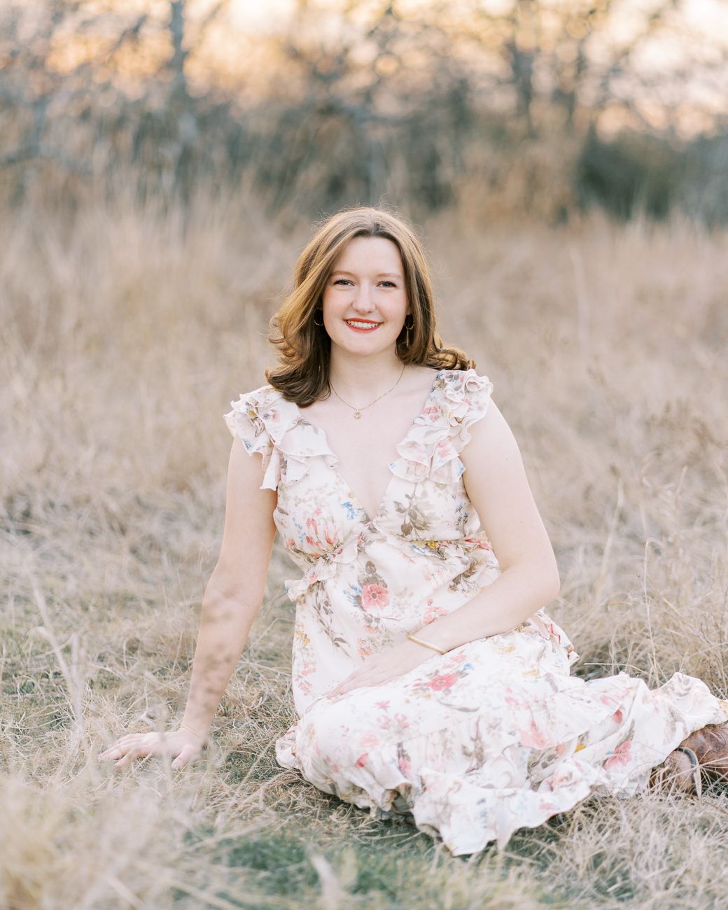A woman in a white floral dress sits gracefully in a sunlit meadow with dried grass and natural surroundings.