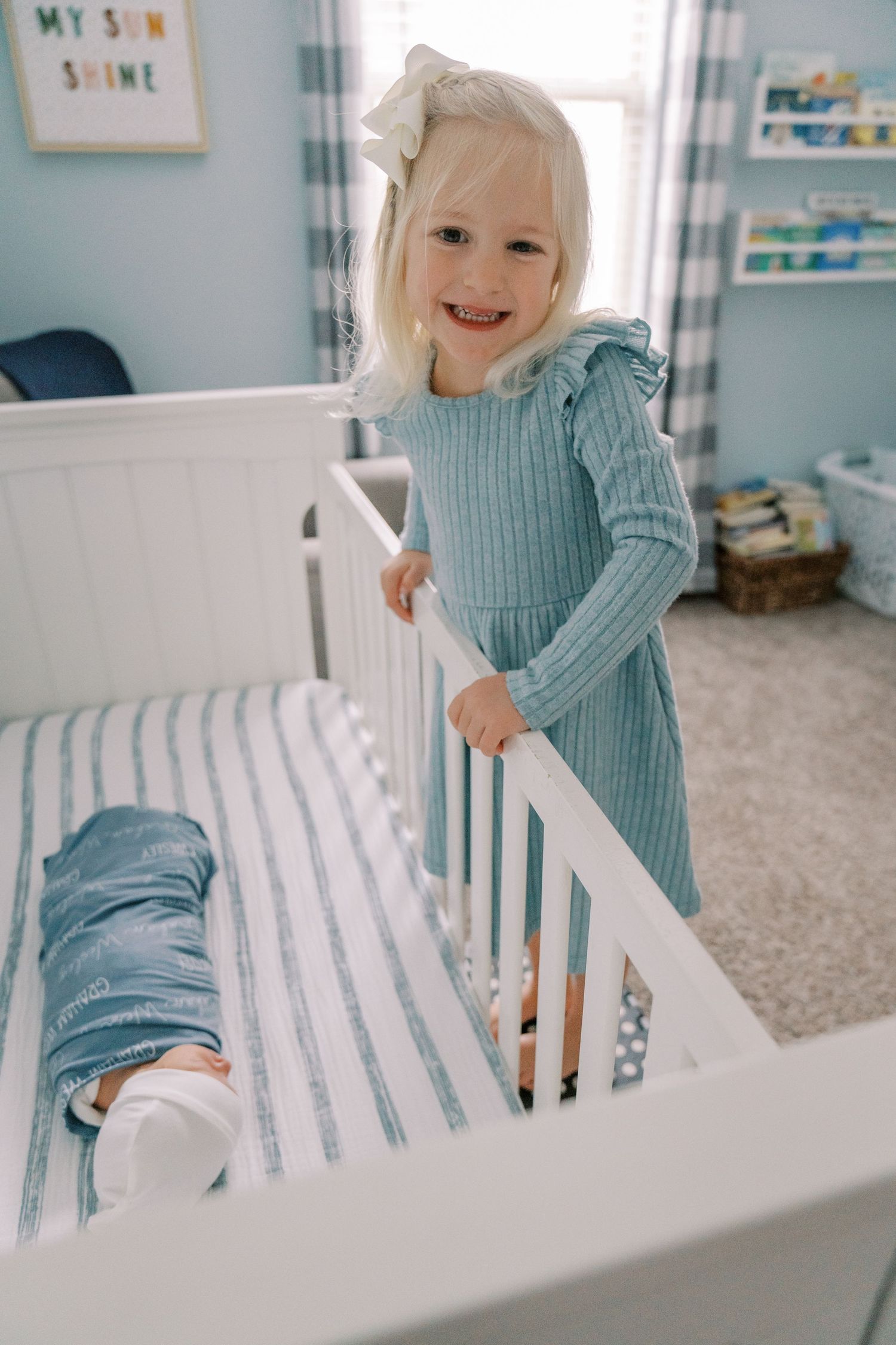 A toddler in a blue sweater plays and smiles while standing in a white crib in a nursery with blue walls.
