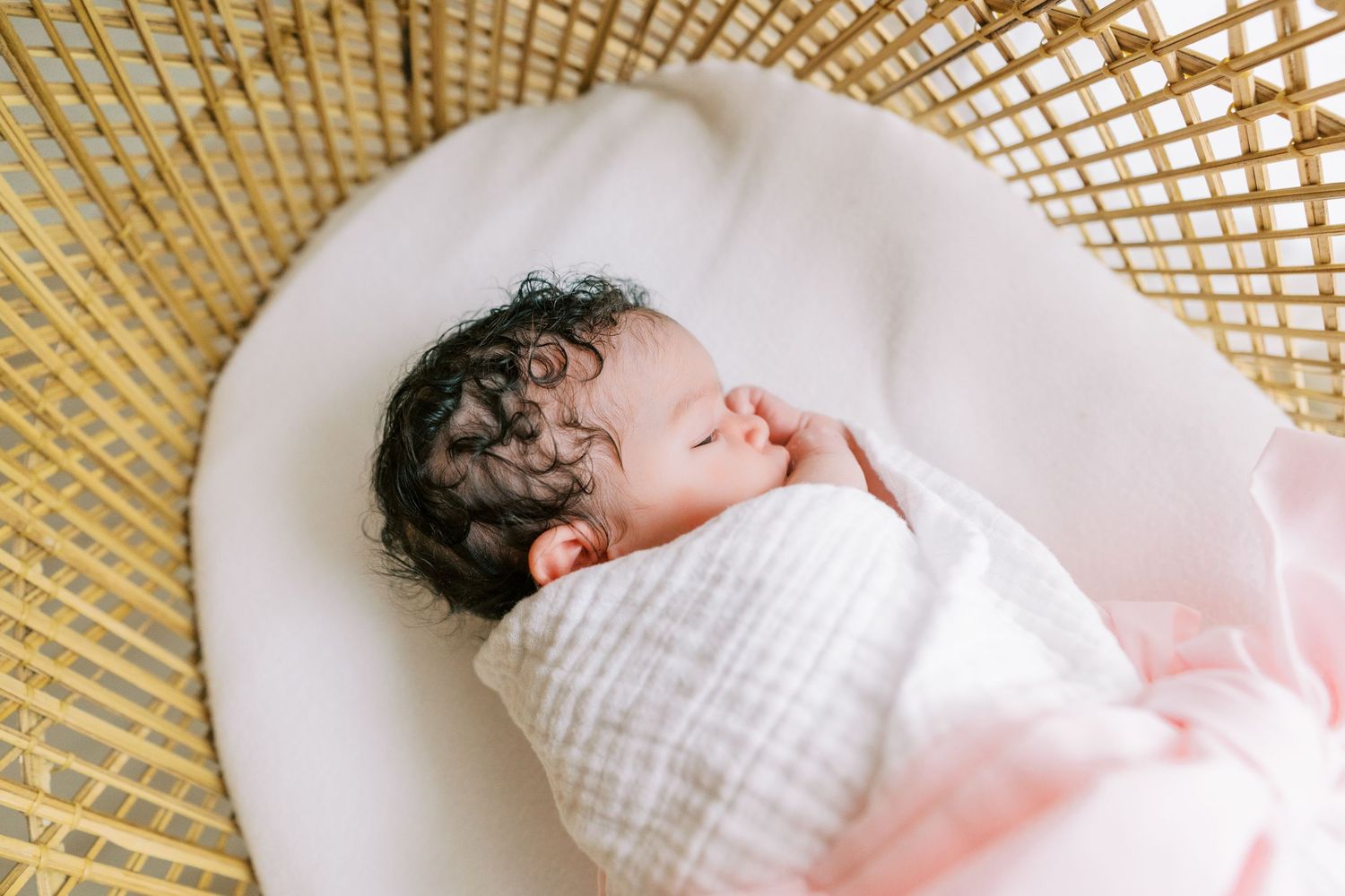Newborn baby with dark curly hair swaddled in white muslin blanket sleeping peacefully in a wicker bassinet.