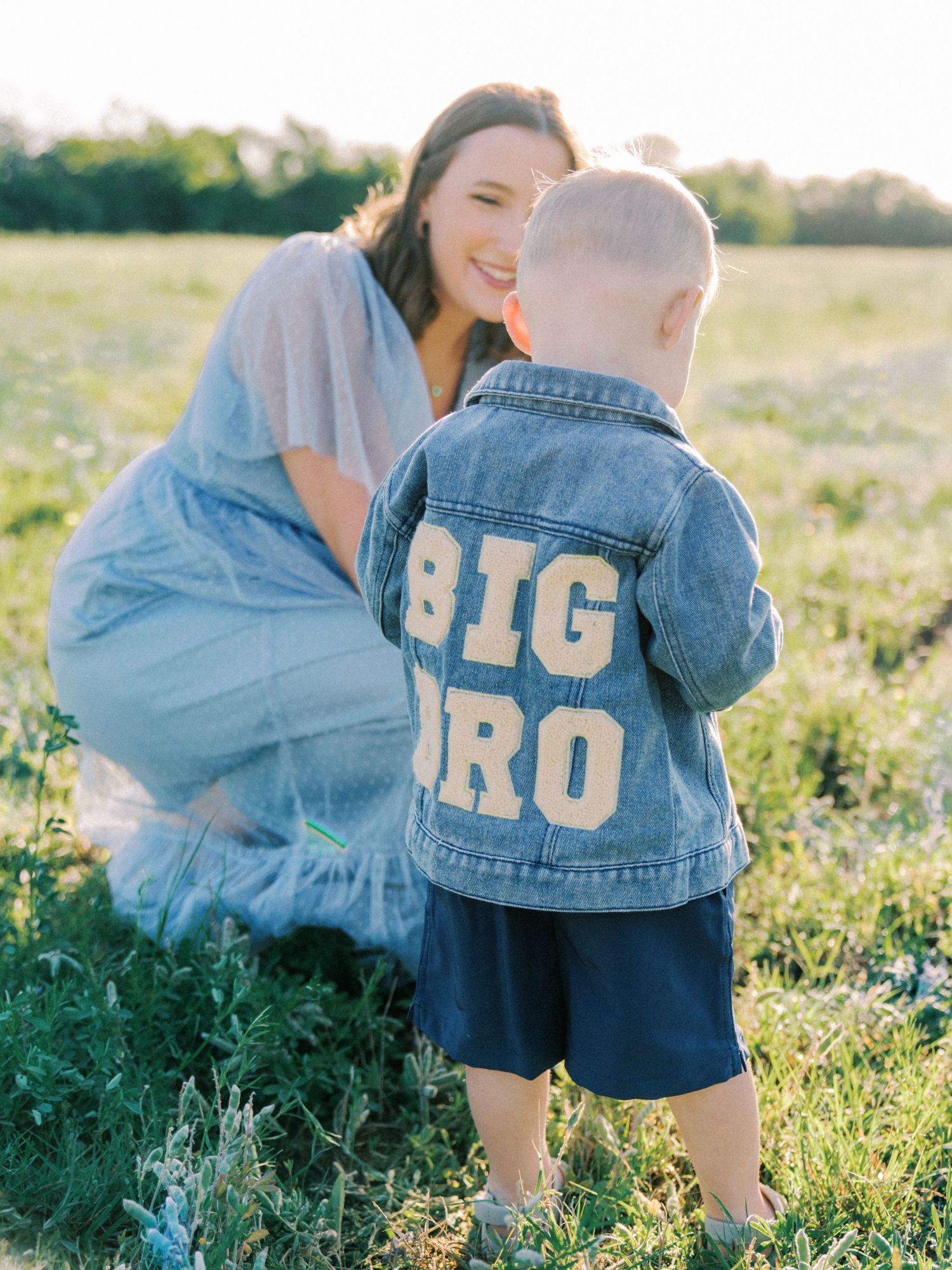 A toddler wearing a denim Big Bro jacket stands with his smiling mother in a sunny green field.