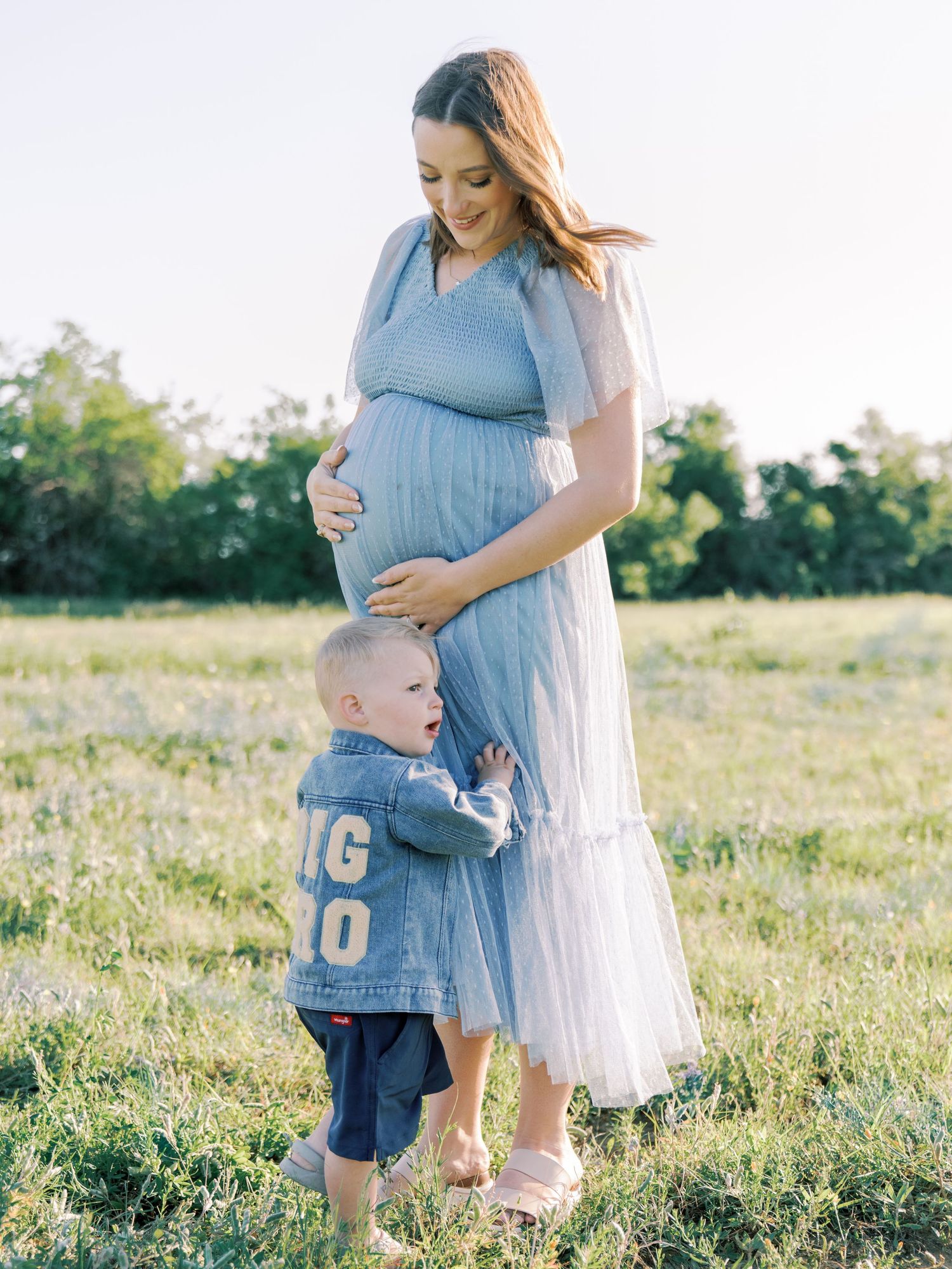 Pregnant woman in blue dress holds belly while toddler in denim jacket stands beside her in sunny field.