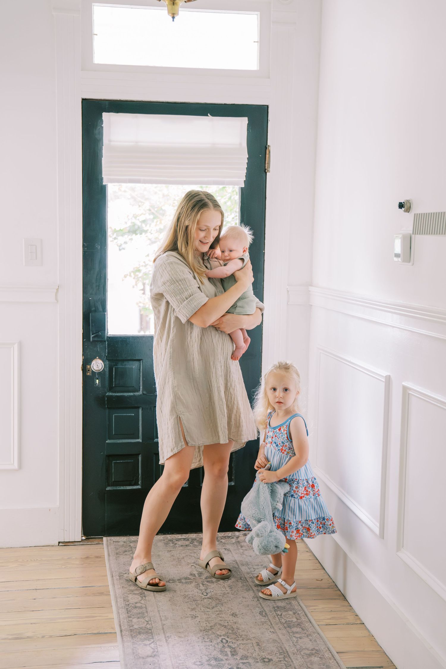 A mother holding her baby near a dark green front door while her young daughter stands beside her in a bright entryway.