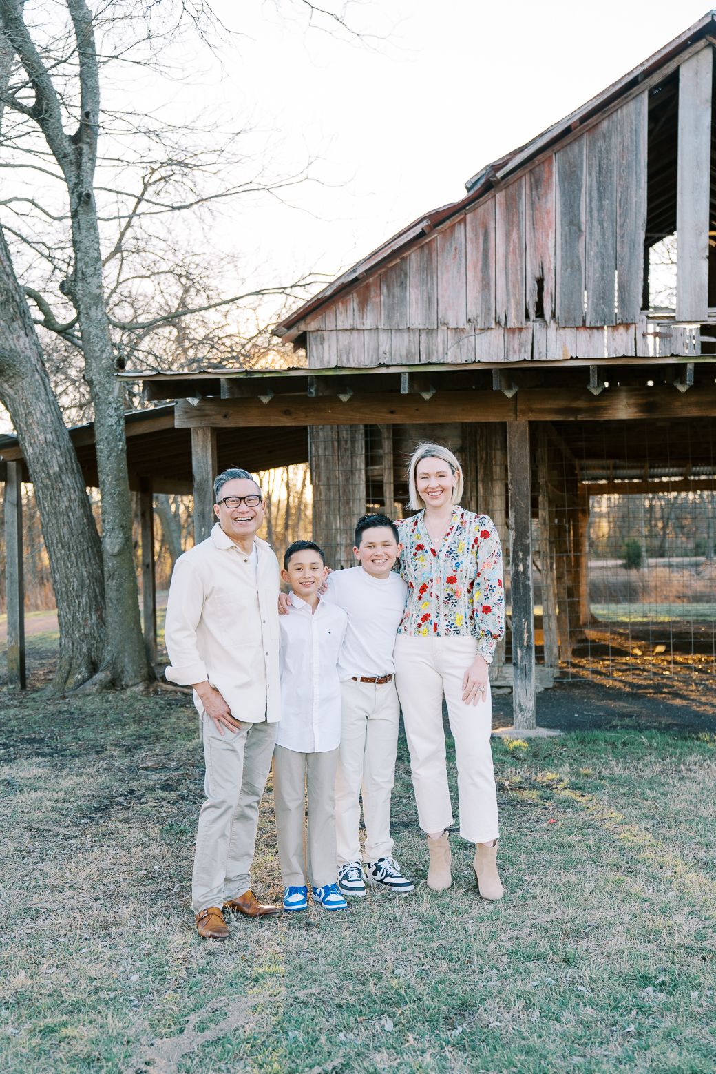 Family of four smiling together outdoors near a rustic wooden barn during golden hour.